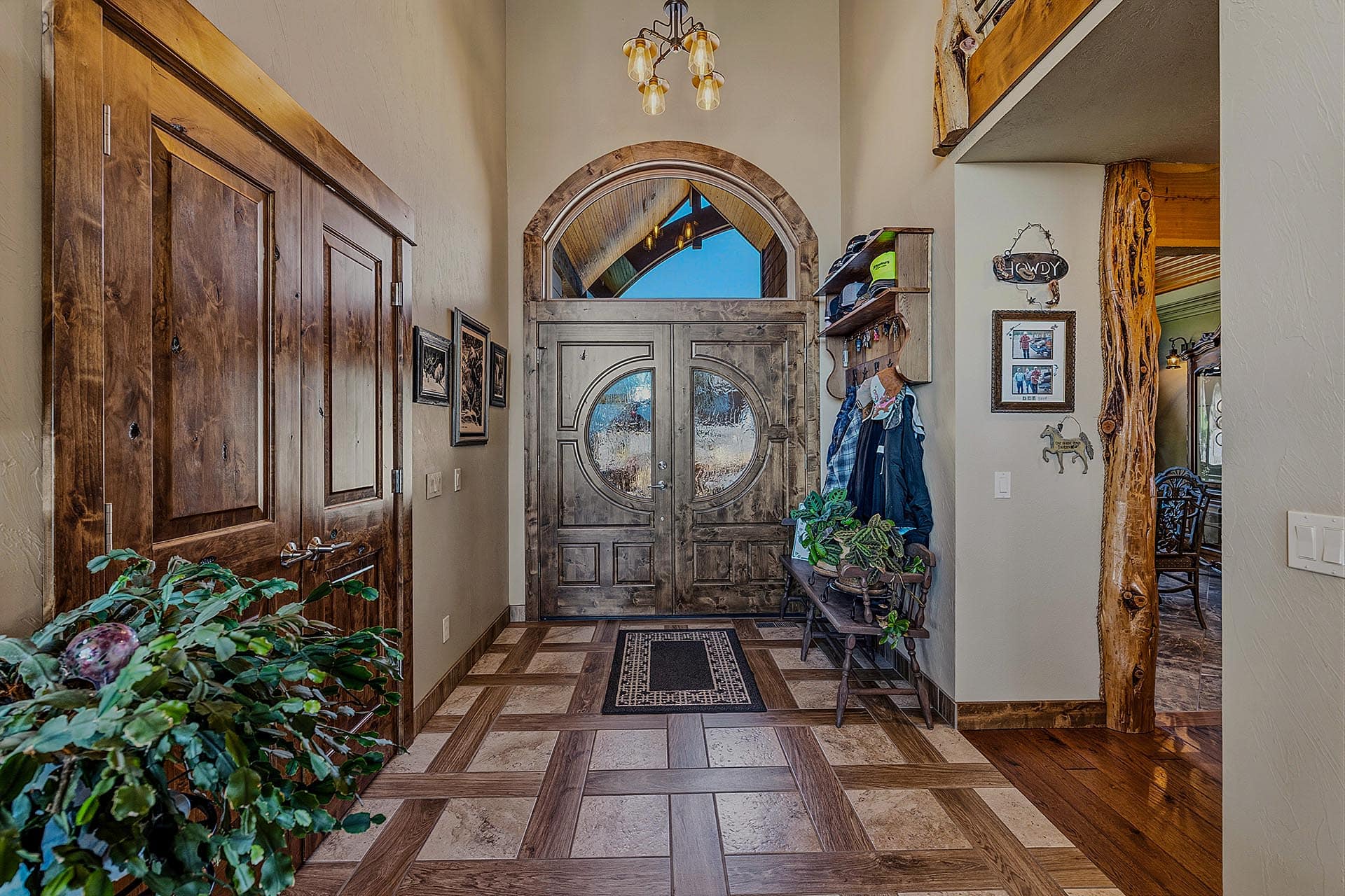 Rustic foyer with heavy wood double doors, arched transom window, and stone accents; coat rack and plants nearby.