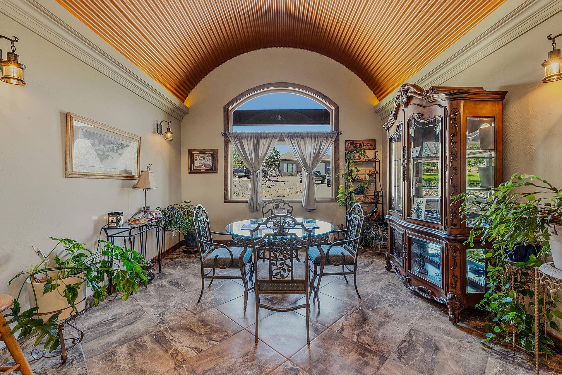 Elegant dining room with a metal-framed glass table, six ornate iron chairs, and a large carved wooden china cabinet beneath a copper arched ceiling.