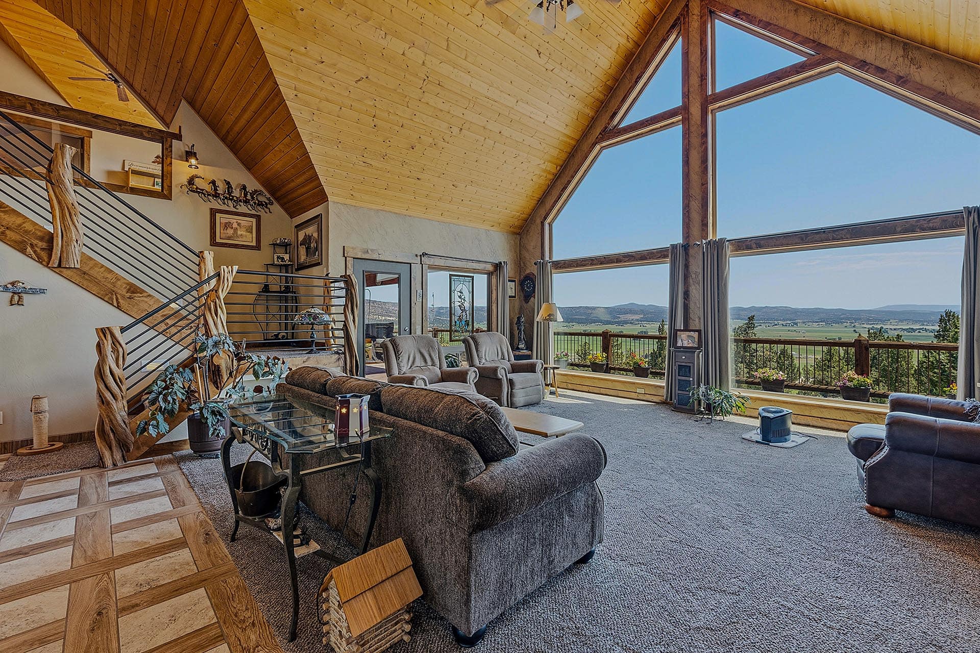 Living room with a vaulted wooden ceiling and large window wall showing a rural landscape; gray recliners and a glass coffee table near a staircase on the left.