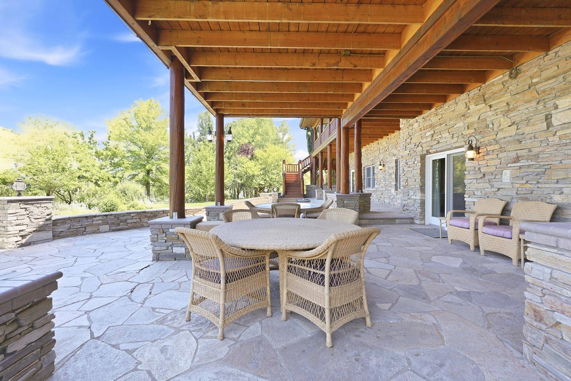 Stone-walled covered patio with wicker tables and chairs under wooden beams, with trees beyond the terrace.