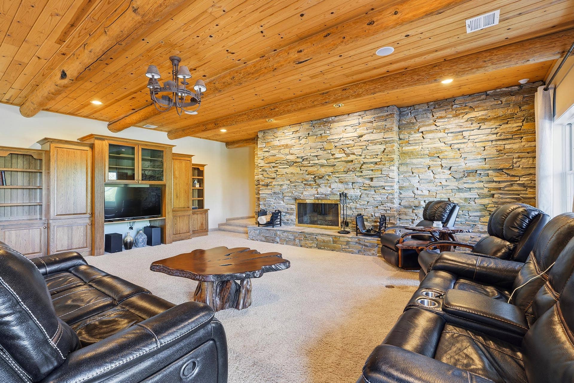 Cozy living room with a stacked stone fireplace, heavy timber ceiling beams, and black leather sofas surrounding a carved wooden coffee table.