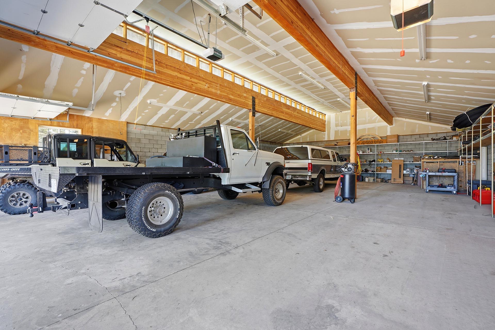 Spacious garage with two pickup trucks parked inside, wooden support beams, and metal shelving along the back wall.