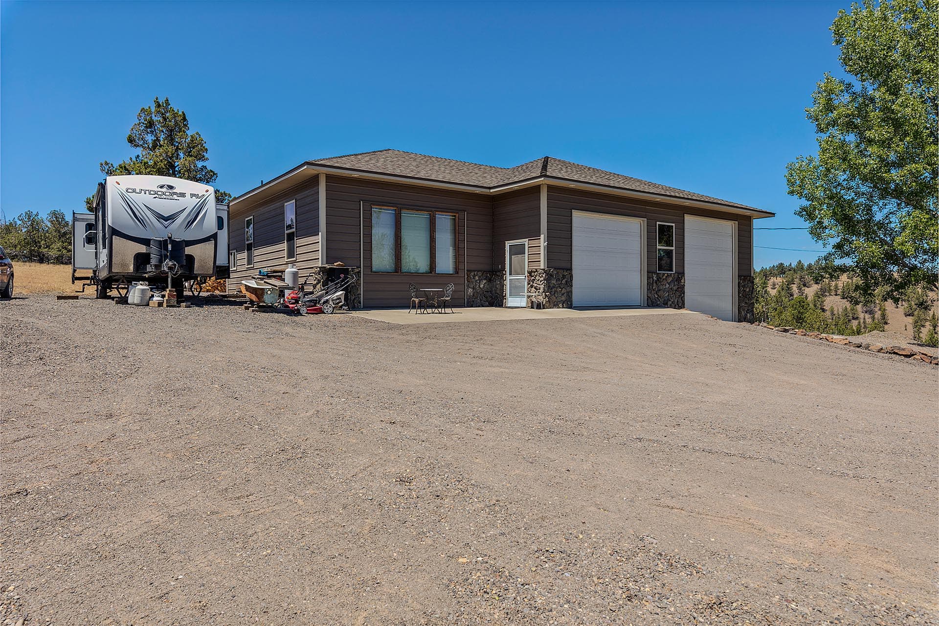 Single-story house with two white garage doors on a gravel driveway; an RV trailer is parked to the left under a clear blue sky.