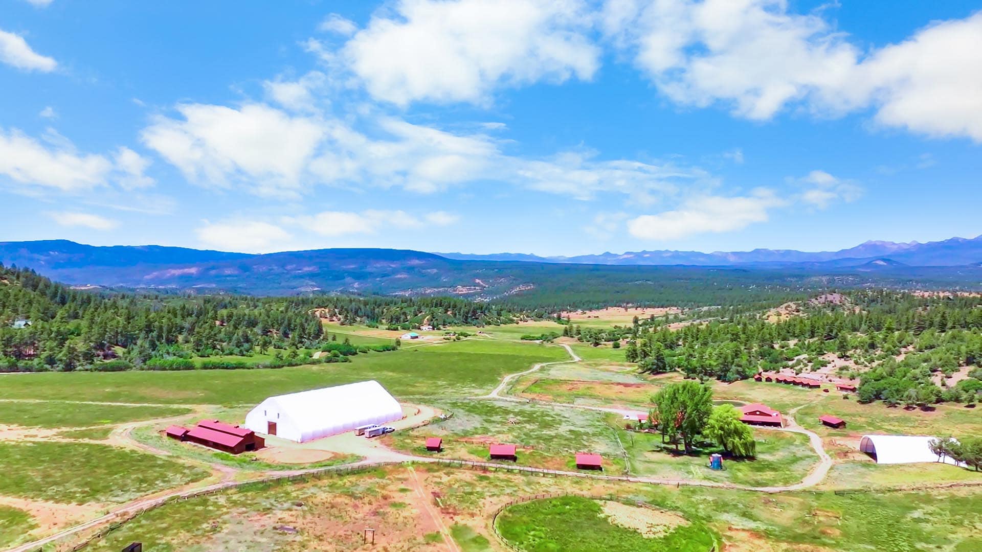 A rural landscape with green fields, scattered red barns, and trees, with rolling hills and distant mountains under a blue sky with clouds.