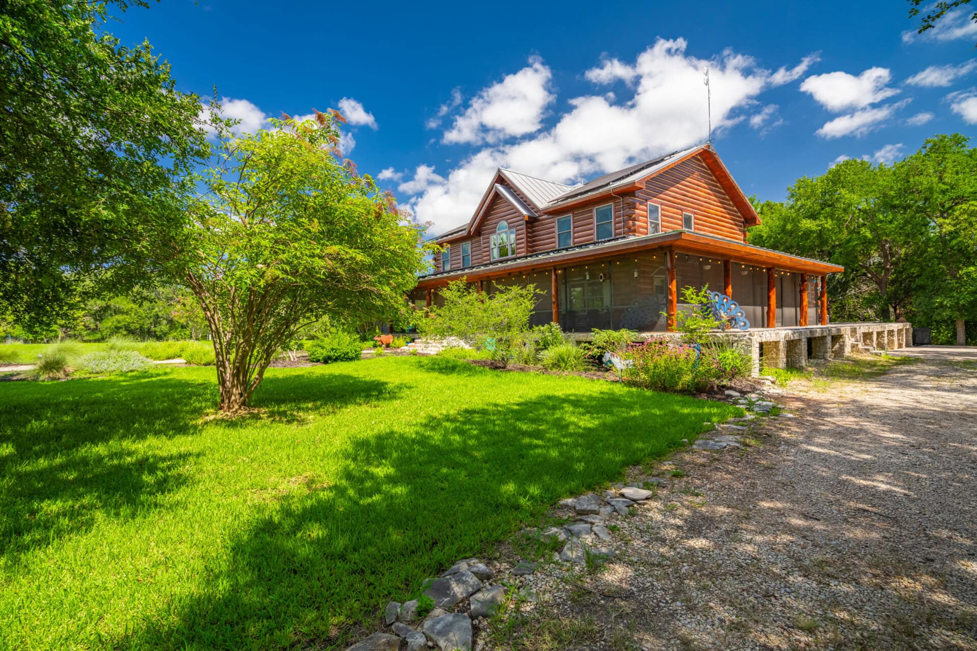 Wooden two-story house with a covered porch, surrounded by green lawn and trees under a blue sky with white clouds