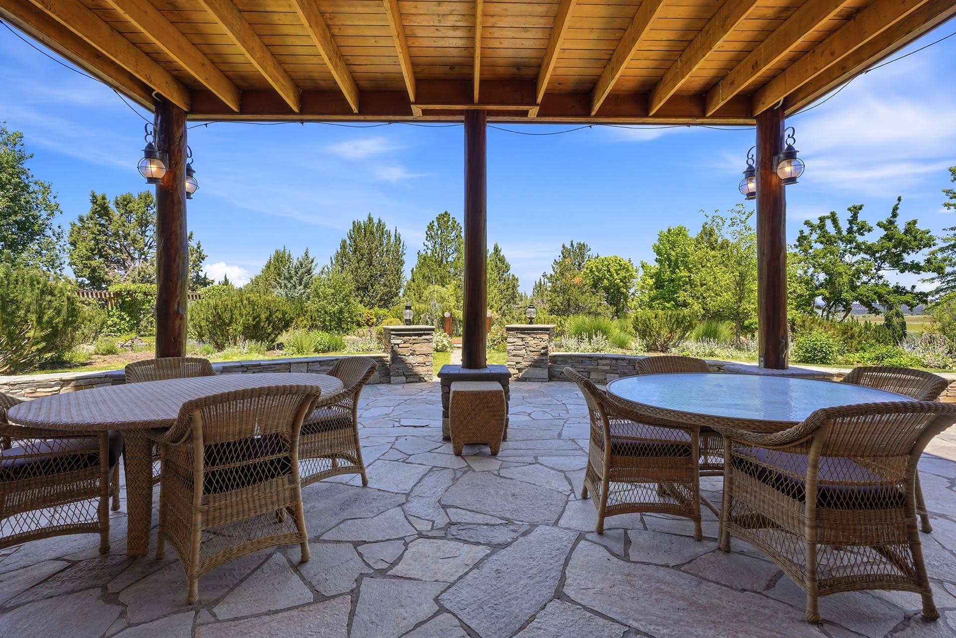 Covered outdoor patio with wicker chairs around a circular glass table, wooden posts, and lanterns, overlooking a garden and blue sky.