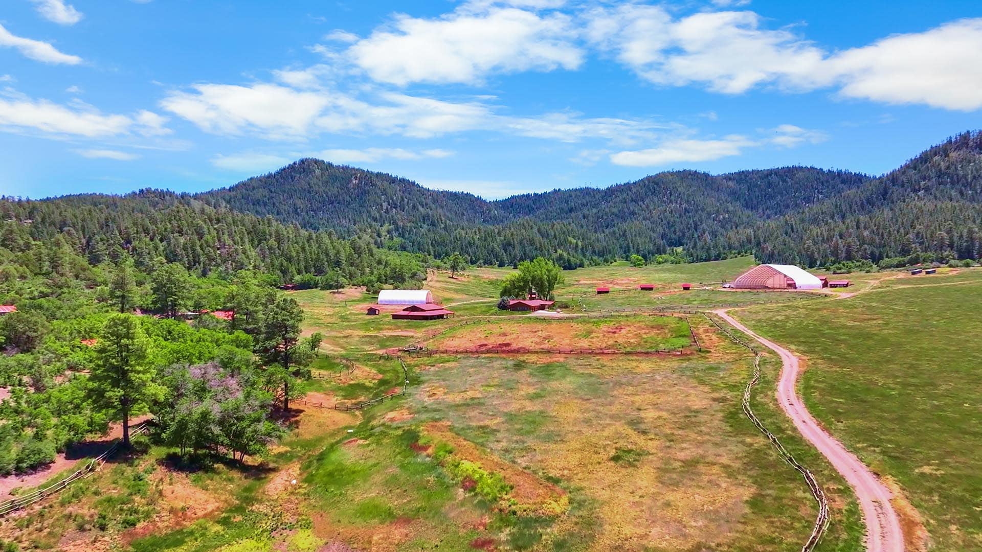 A rural farmstead in a valley with rolling hills, red barns, and a winding dirt road under a bright blue sky.