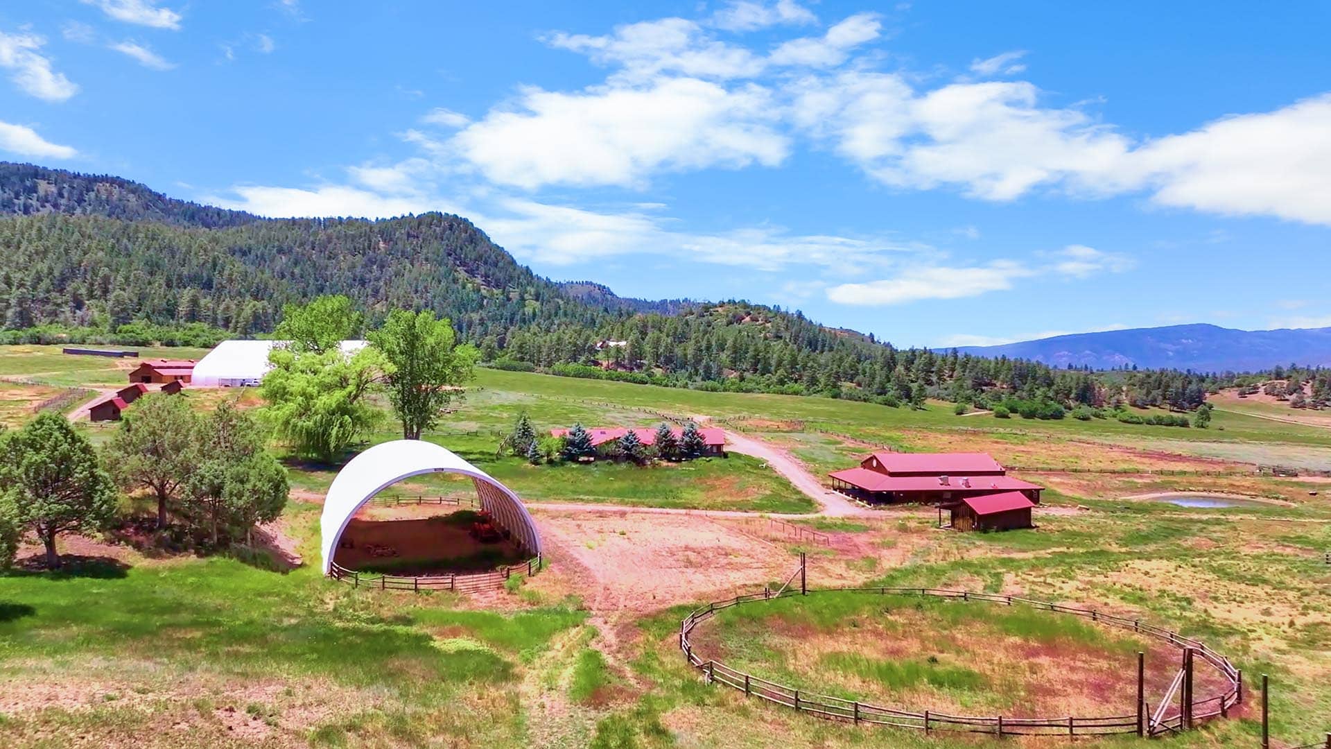 Rural farm landscape with red barns, a white curved shelter, green pastures, and forested hills under a blue sky.