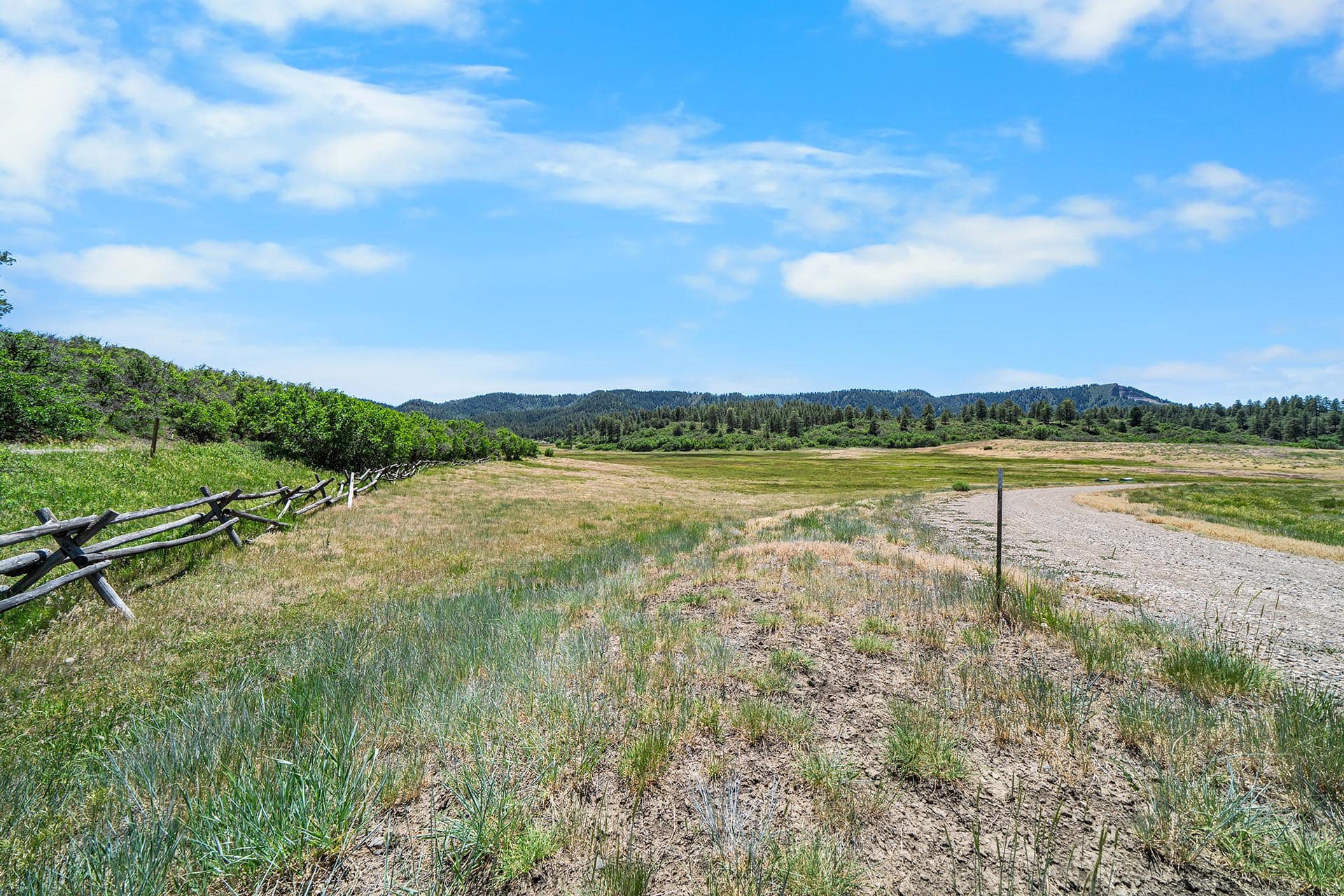 Open grassy field with a wooden split-rail fence along the left, rolling hills in the distance under a blue sky with wispy clouds.