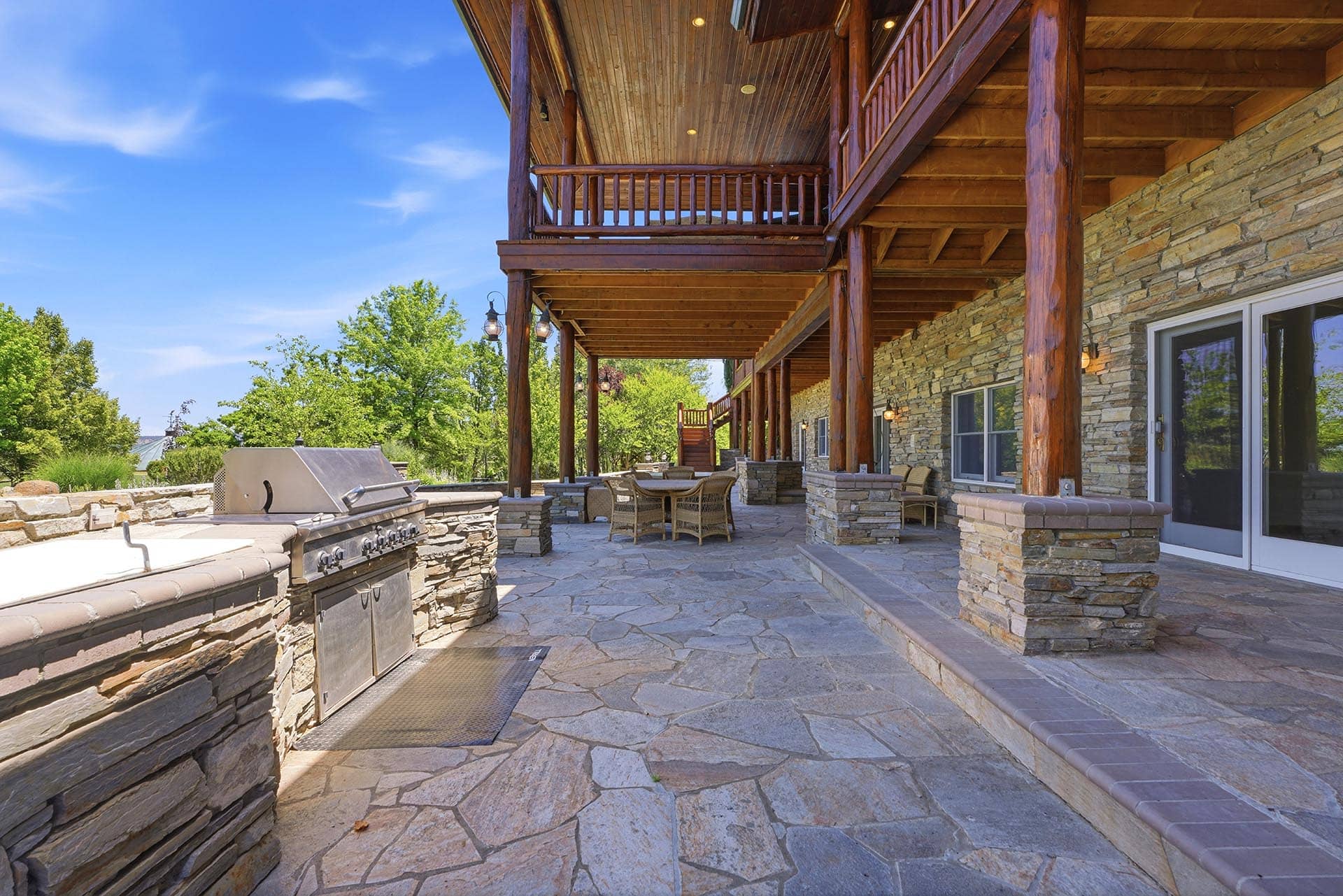 Stone-paved patio with a built-in grill, wooden log columns, and stone veneer walls; outdoor seating under a wooden balcony on a sunny day.
