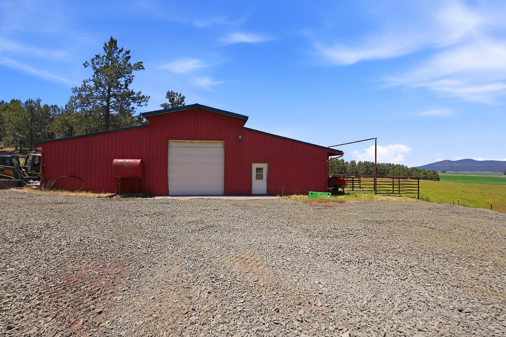 Red barn with a white garage door on a gravel lot, blue sky overhead and distant hills to the right, farm equipment on the left.