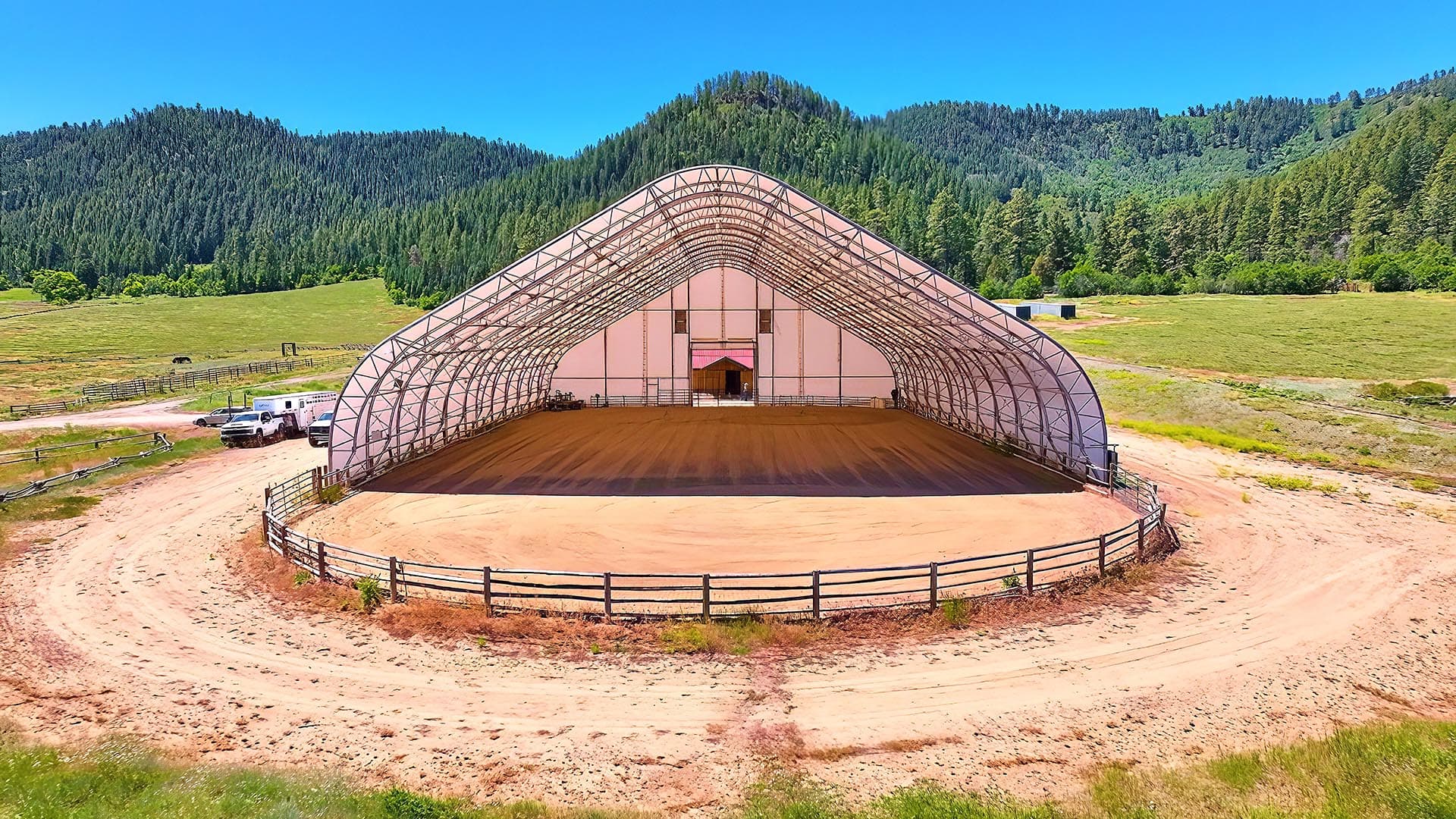 Open-sided, dome-shaped barn with a dirt arena inside, set in a rural field with mountains in the background and a few vehicles nearby