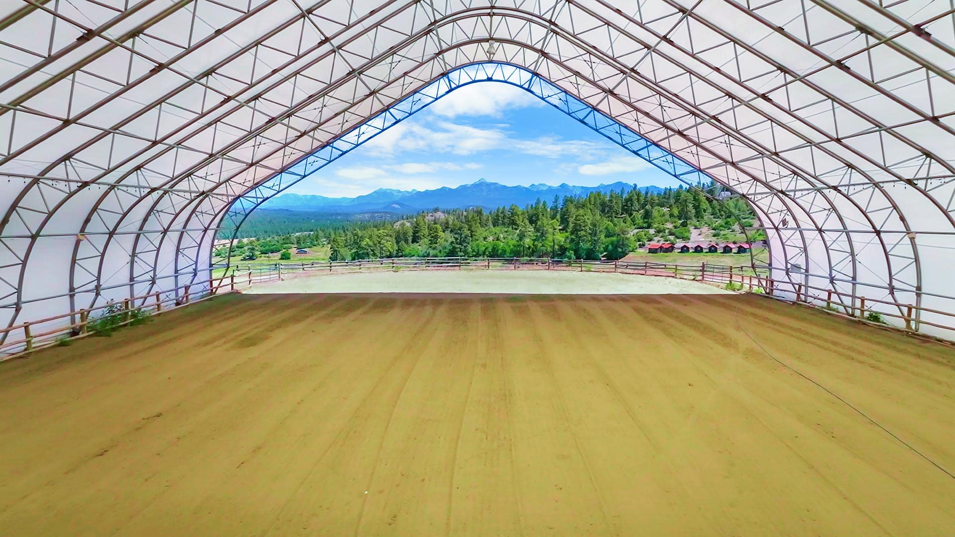 Interior of a large curved metal-frame shelter with a wooden floor, opening to a mountain landscape with trees and a fence beyond