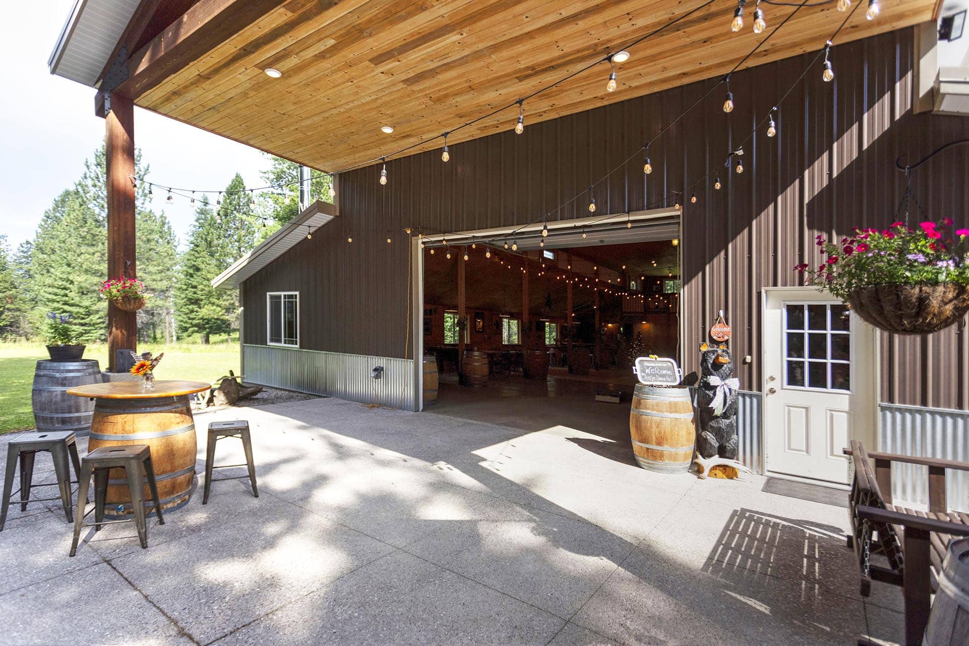 Rustic barn patio with string lights, wooden barrels as tables, and hanging flower baskets outside the open doorway.