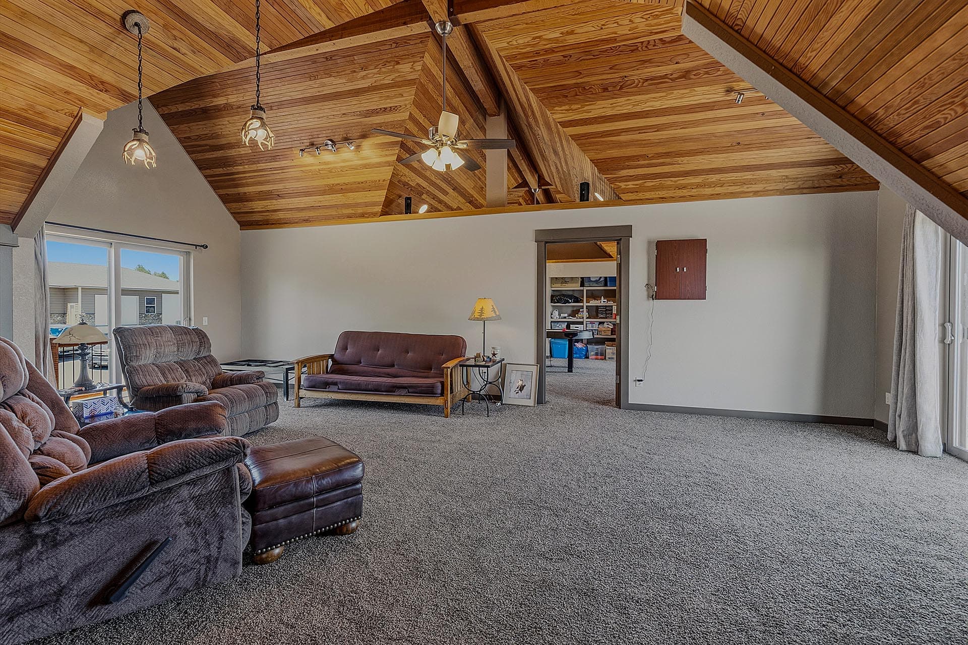 Living room with a dramatic wood vaulted ceiling, brown seating, and a carpeted floor; sliding glass doors open to outdoors on the left.