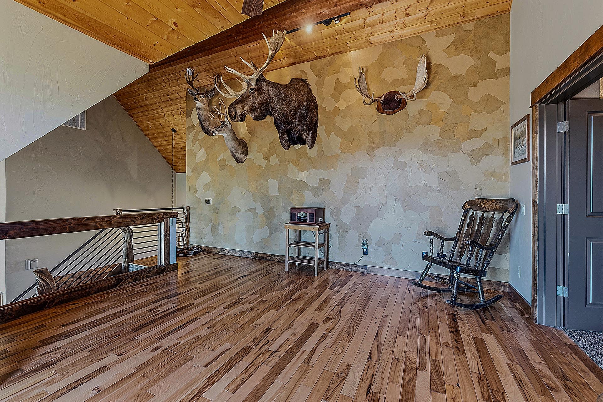 Loft-style living area with three mounted animal heads on a stone-pattern wall, wood ceiling, and wooden floors with rocking chairs nearby.