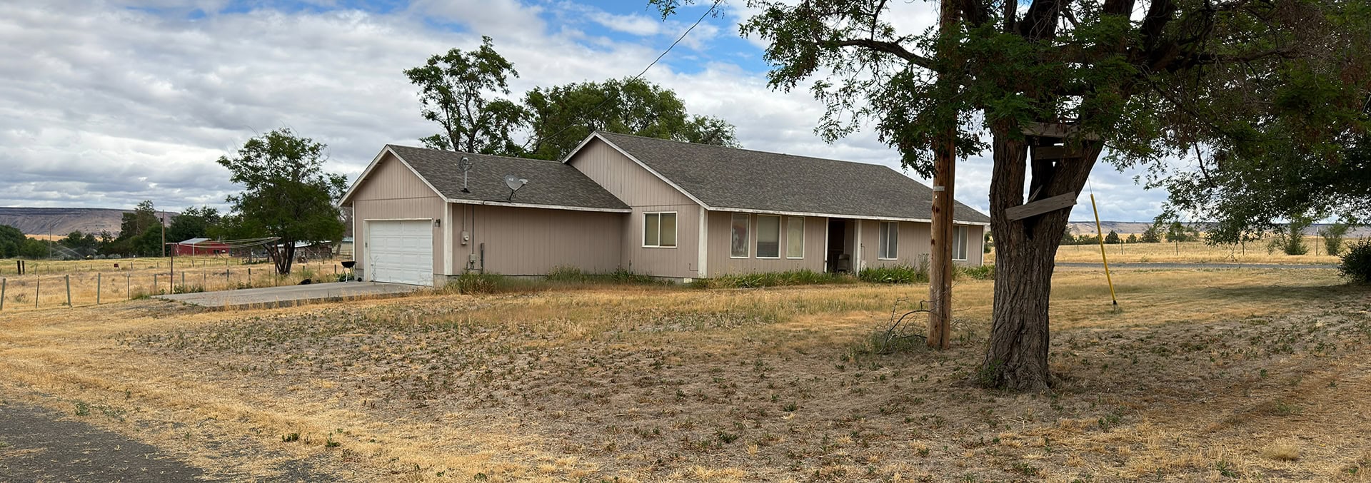 Rural single-story house with a detached garage on a dry grassy yard under a cloudy sky; a large tree stands to the right with a wooden platform attached.