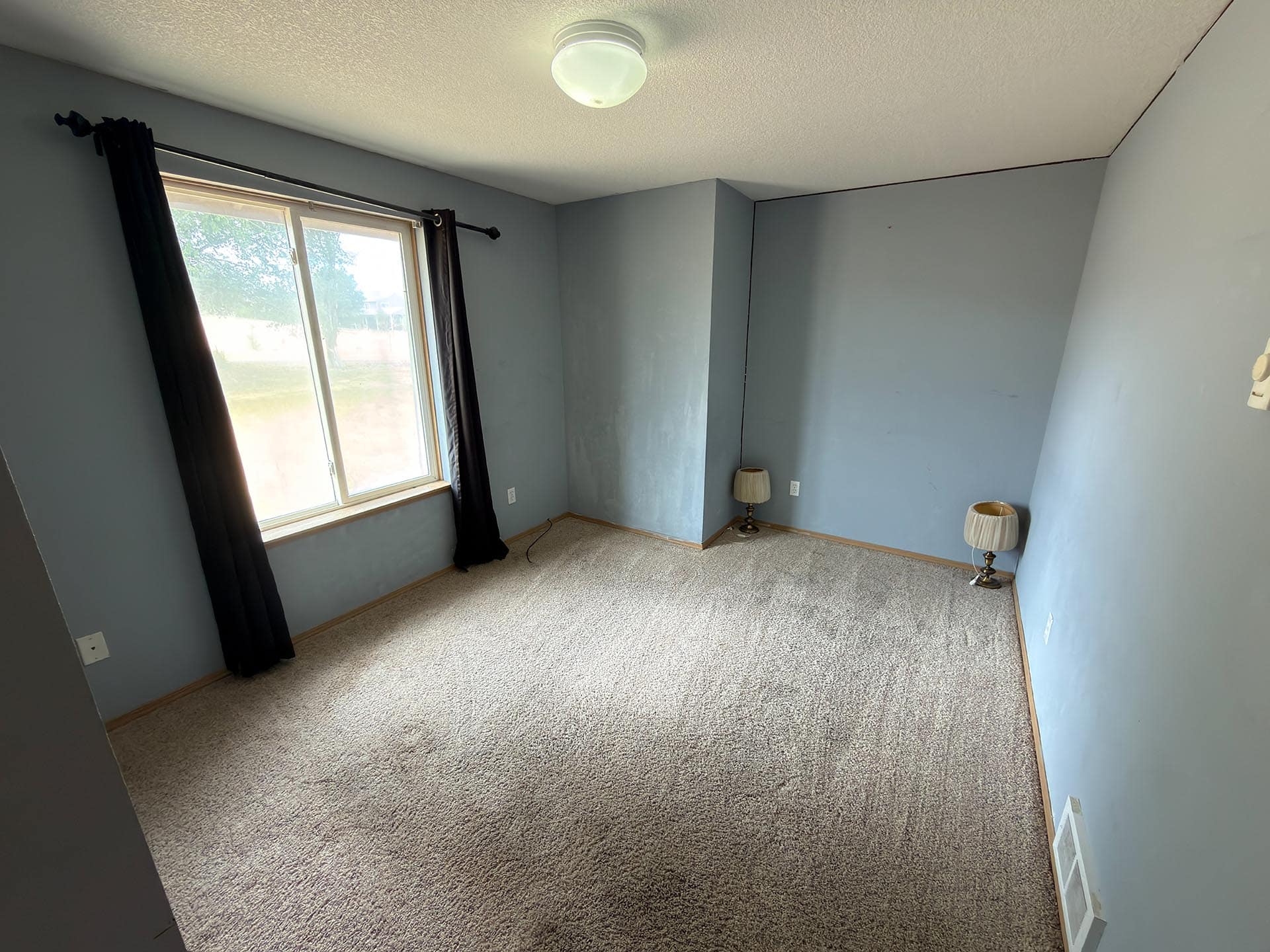 Empty blue-gray bedroom with a window on the left, dark curtains, and two small lamps on the floor by the walls.