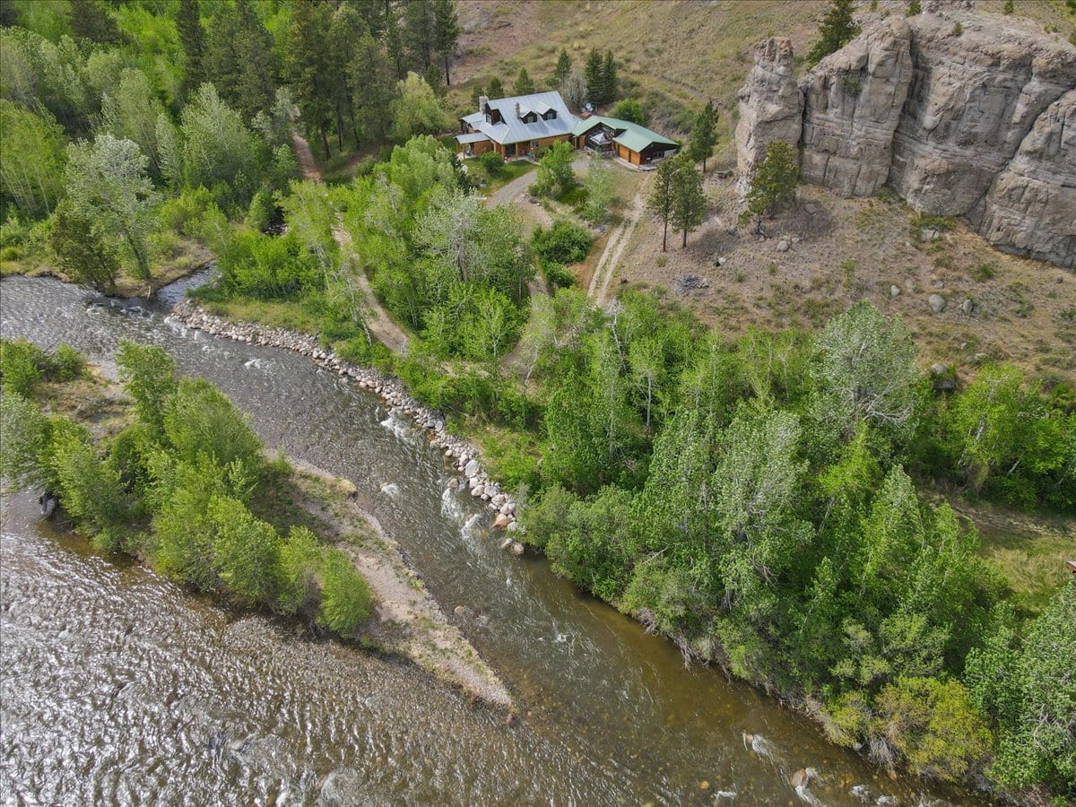 Aerial view of a rustic house with a metal roof beside a winding river, surrounded by trees and rocky cliffs.