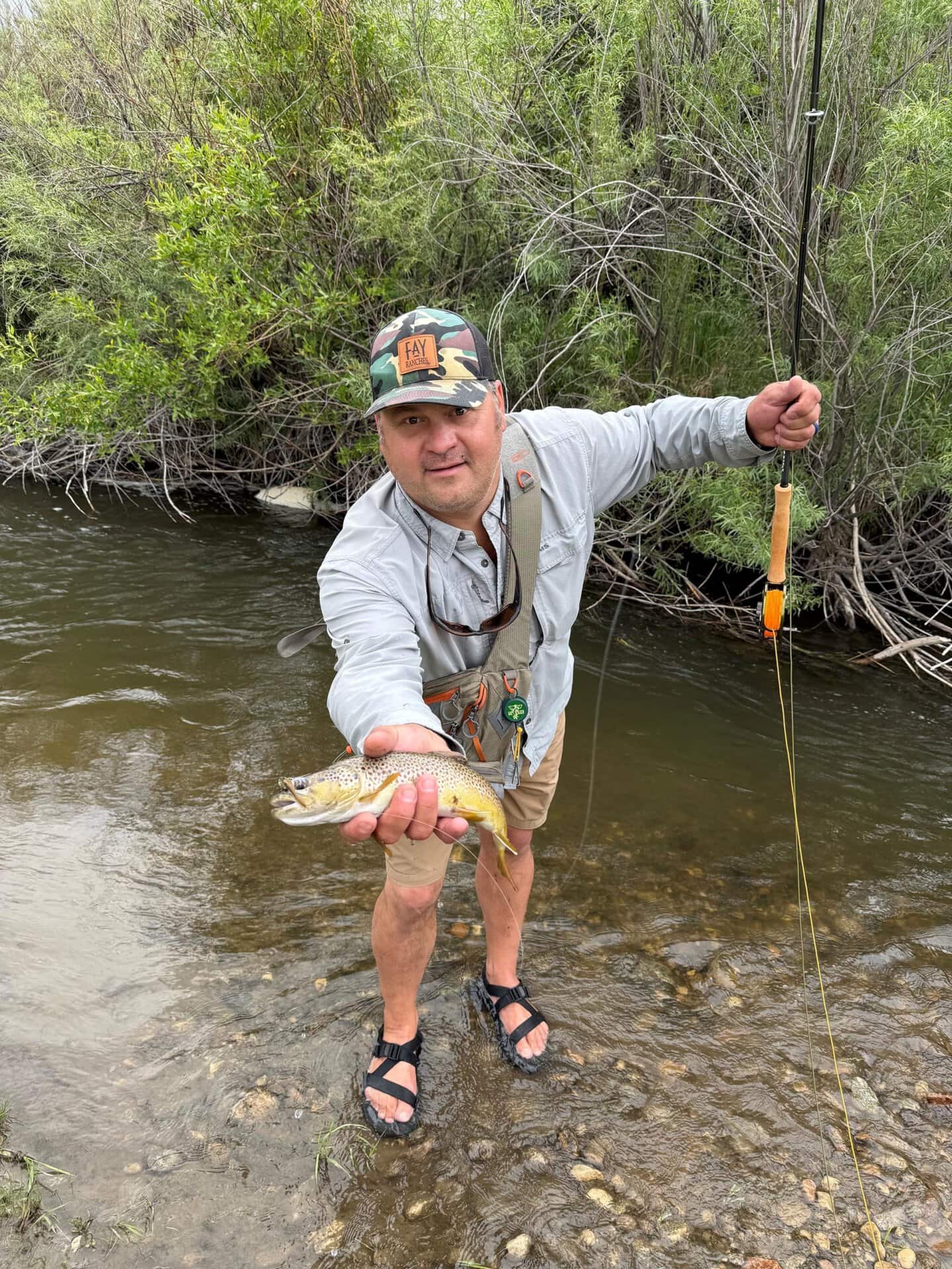 Man wearing a camouflage hat and light shirt poses in a shallow river, holding up a small fish he just caught with fishing gear visible.