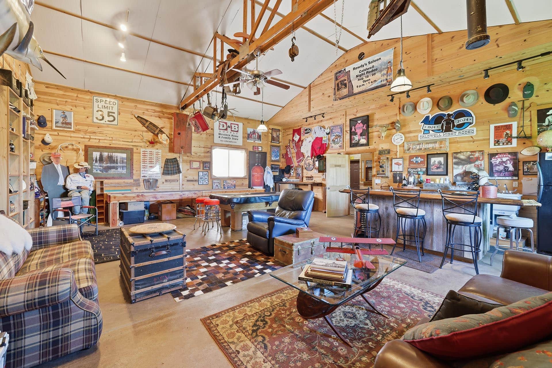 Cozy rustic man cave/bar with wood walls, vintage signs, hats, and a seating area by the long counter on the right.