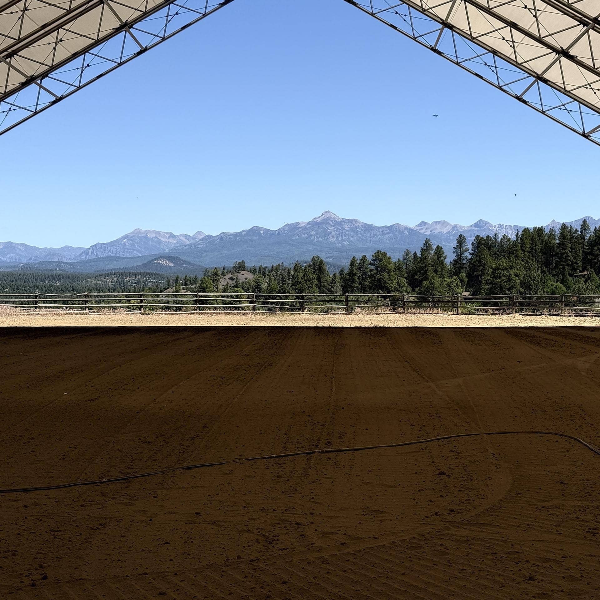 Open-air arena with sandy ground and a wooden fence, mountains in the distance under a clear blue sky, metal frame overhead.