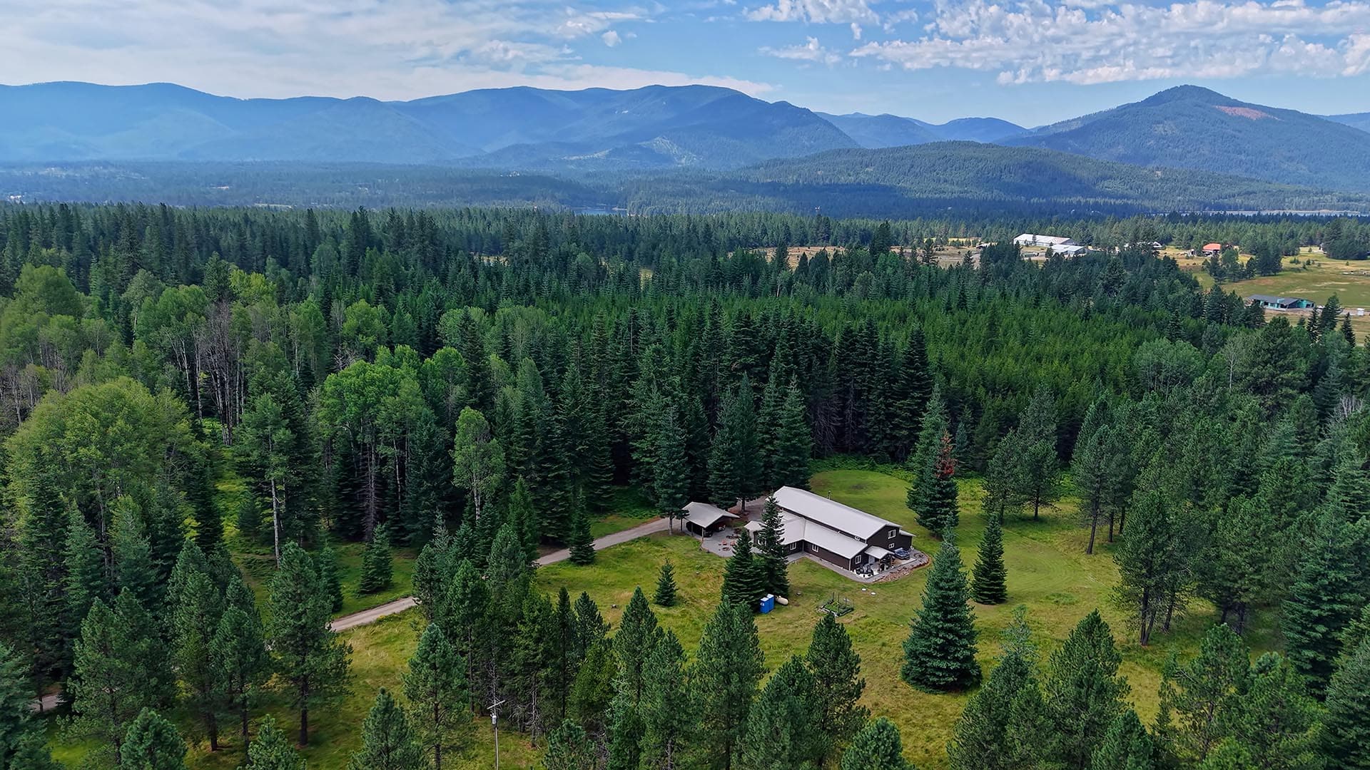 Aerial view of a dense evergreen forest with a large farmhouse complex at center, mountains in the distance.