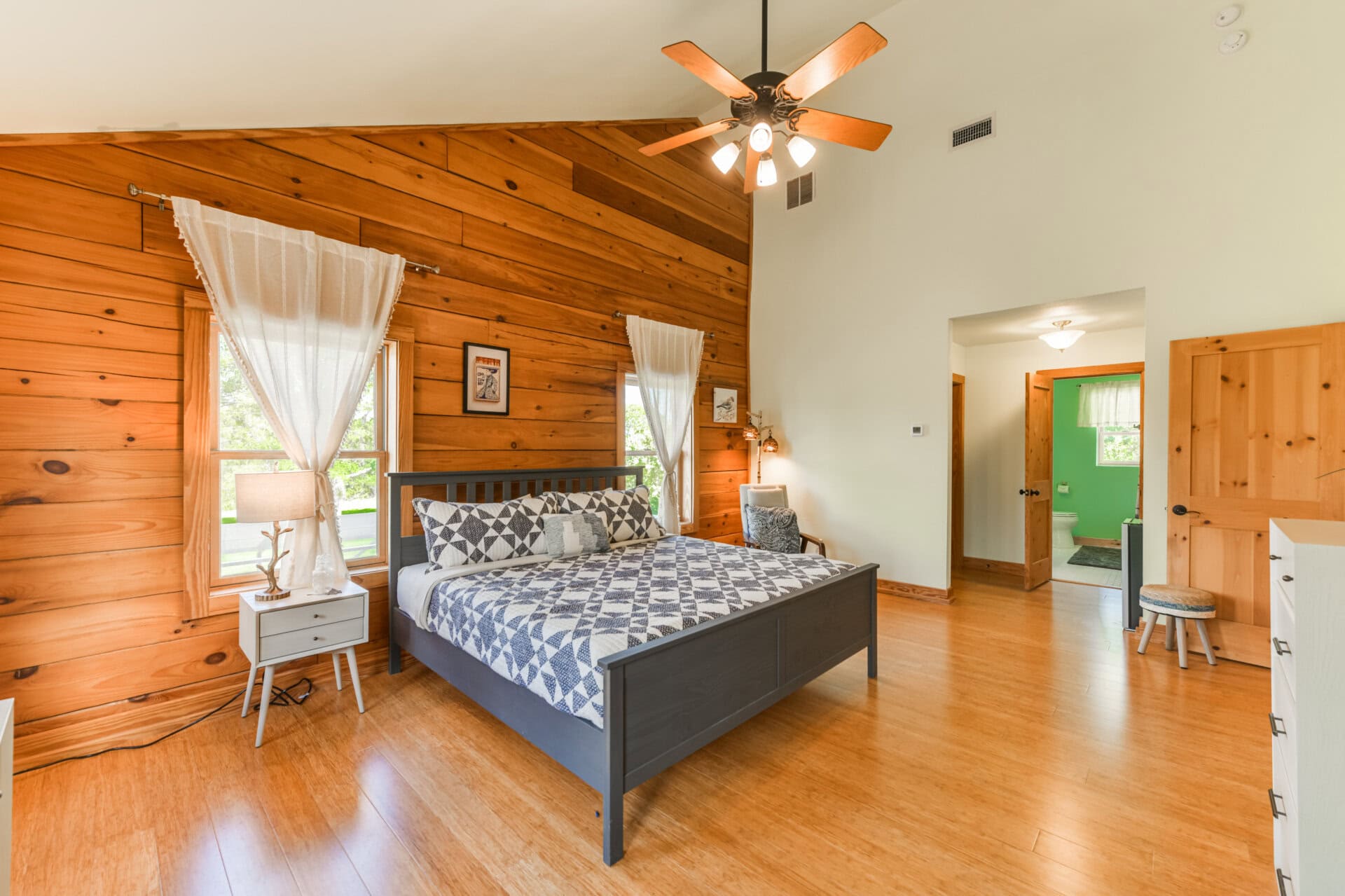 Bedroom with a dark gray bed, geometric quilt, white nightstands, and a warm wood-paneled wall; ceiling fan above.