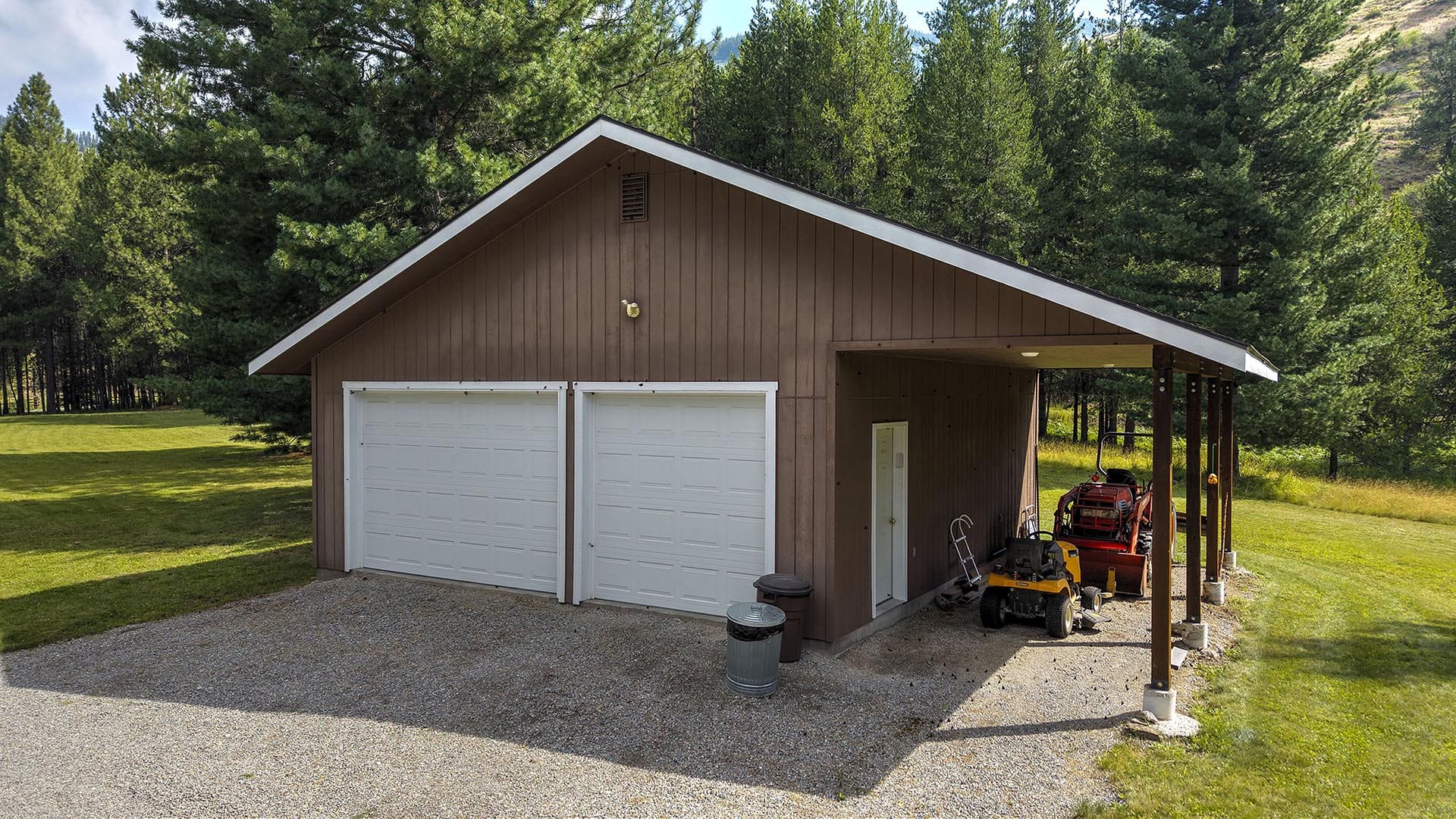 Brown wooden garage with two white doors, side covered area housing lawn equipment among trees in a grassy yard.