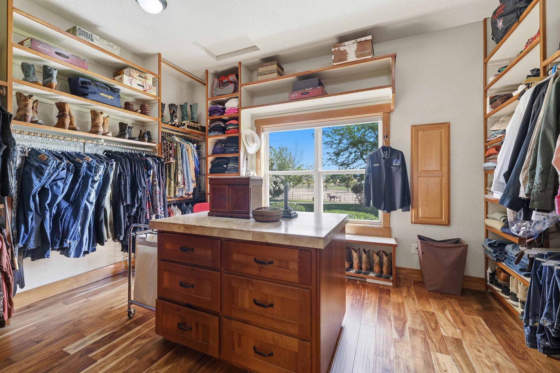 Bright walk-in closet with wooden shelves, jeans and boots on the left, shirts on racks, and a central wooden island vanity under a large window.