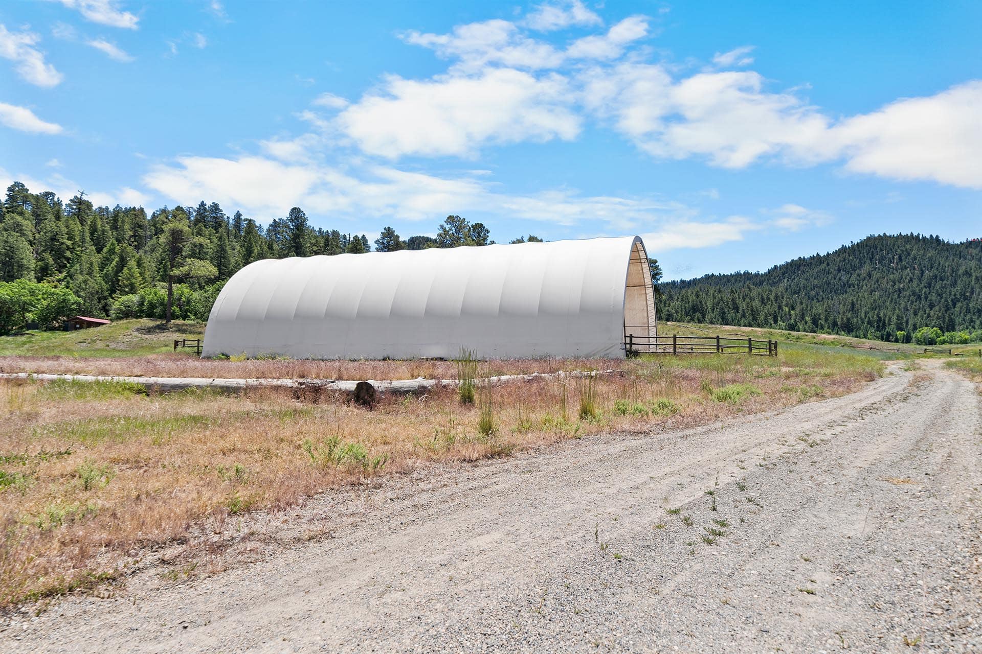 White arched storage shelter in a rural field under a blue sky with pine trees and distant hills.