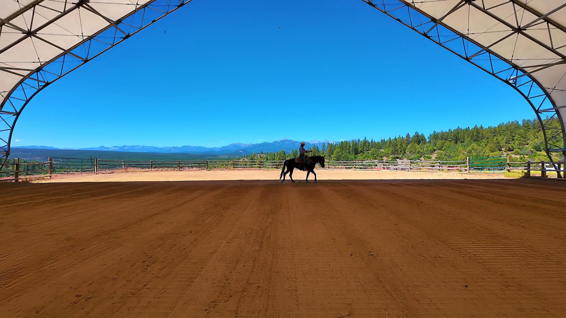 Rider on a dark horse walks in a sandy riding arena with mountains in the distance under a blue sky