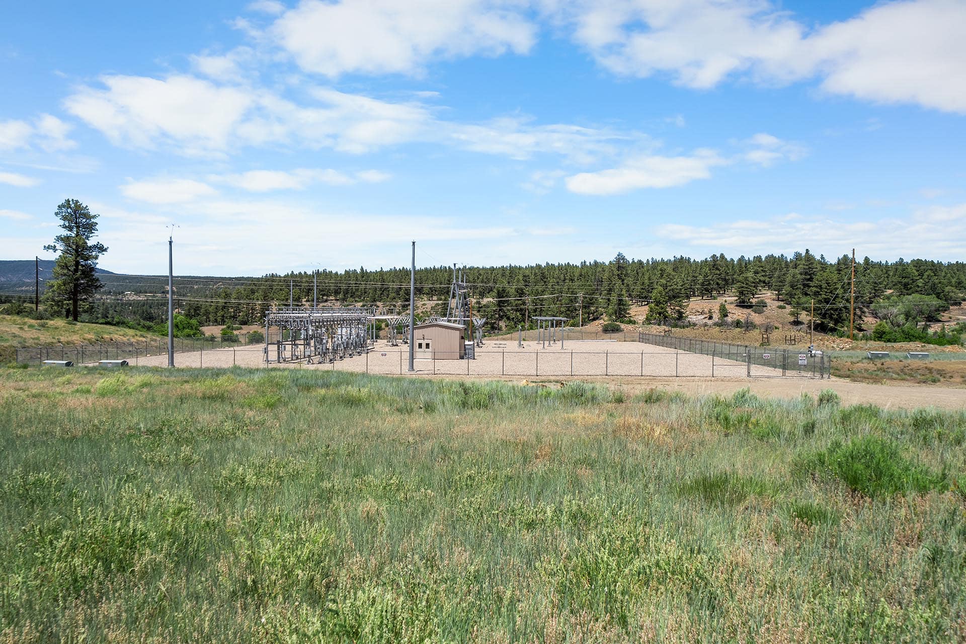 Rural electrical substation with metal structures and power lines inside a fenced area, under a blue sky with scattered clouds.
