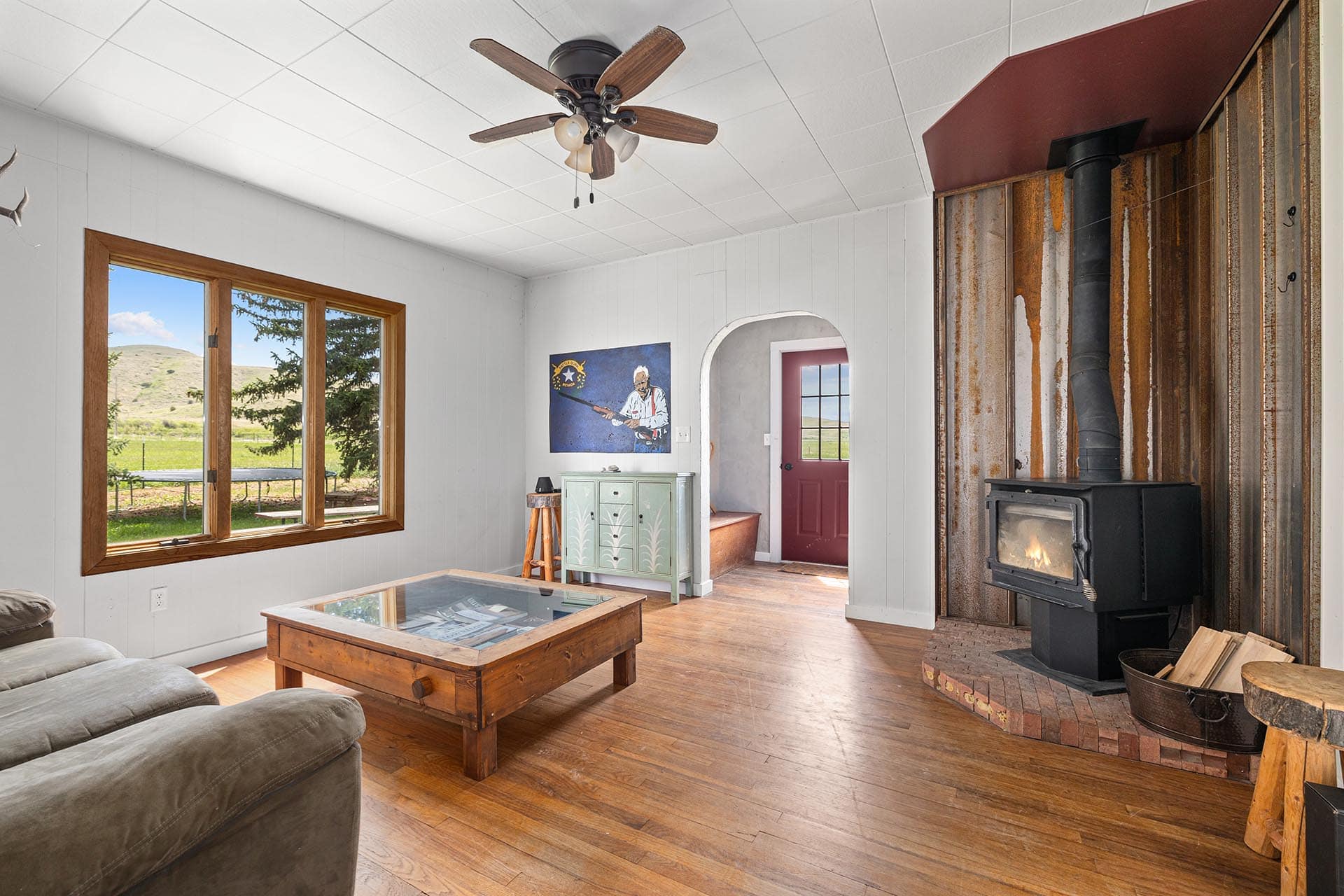 Cozy living room with a wood stove on a brick hearth and rusted wall paneling, facing a large window to a rural landscape.