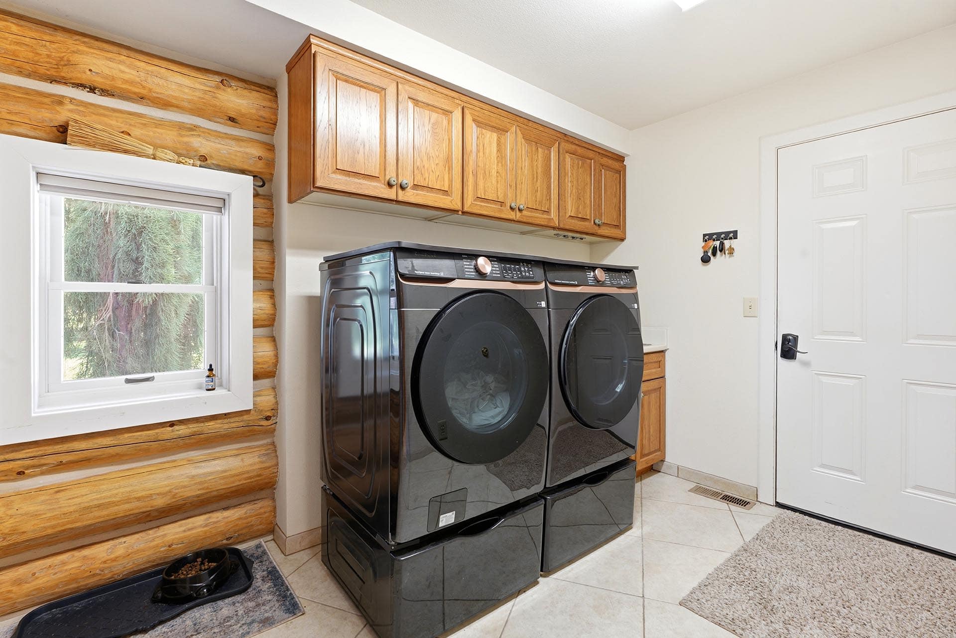 Stacked dark gray front-loading washer and dryer beneath wooden cabinets in a laundry room, with a window on a log wall to the left.