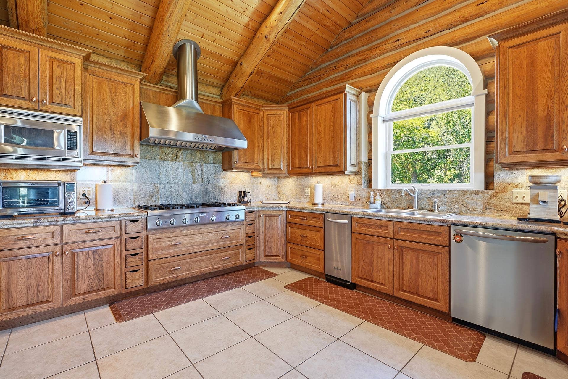 Rustic kitchen with wooden cabinets, a stainless steel range and hood, and a large arched window revealing greenery.