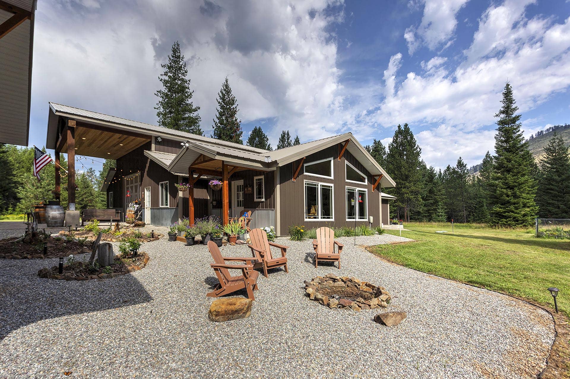 Wooden lodge-style building with a covered porch, gravel courtyard, and several Adirondack chairs arranged around a stone fire pit outdoors.