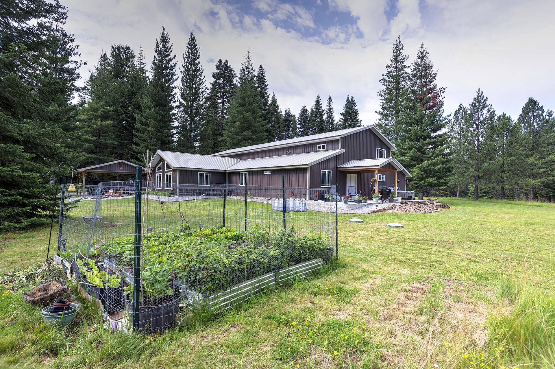 Brown house with a metal roof sits in a spacious yard, bordered by tall evergreen trees; foreground shows a fenced vegetable garden.