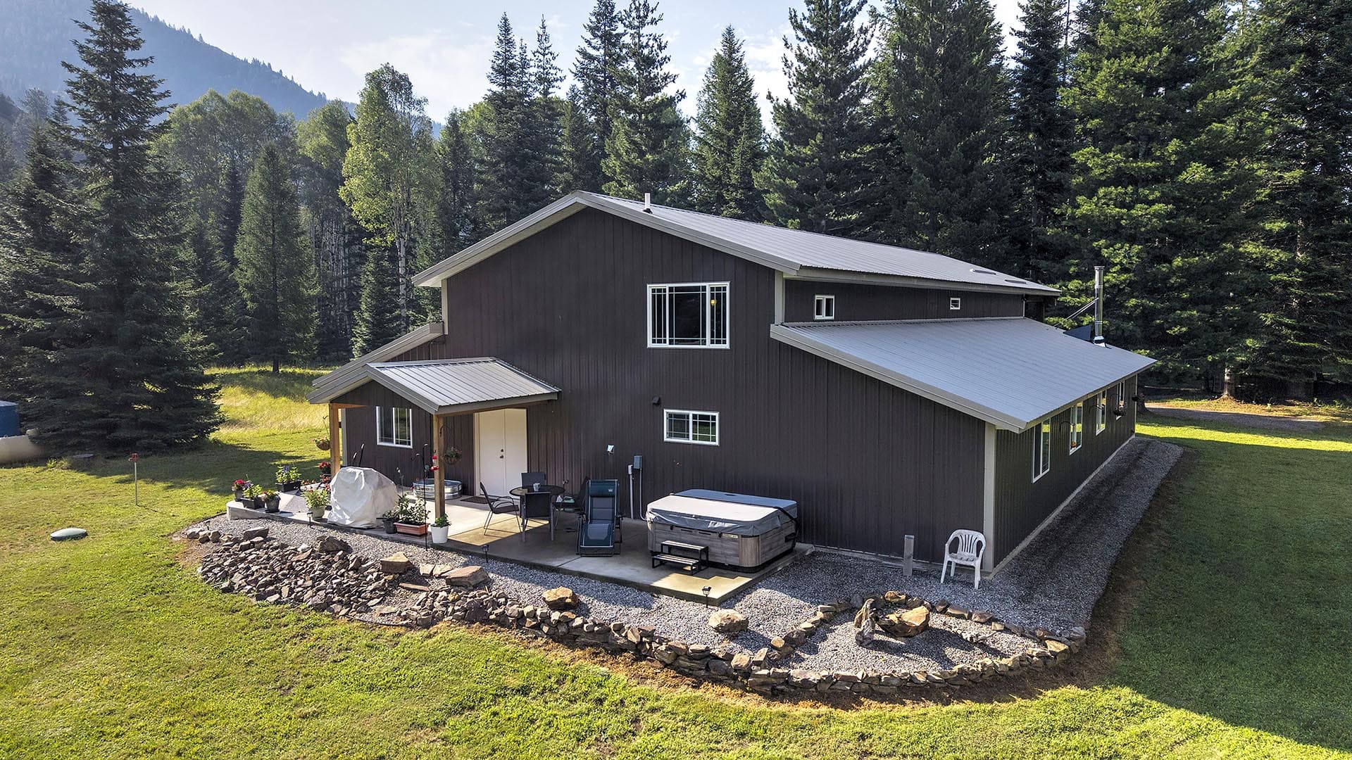 A dark gray house with a metal roof in a forested yard, featuring a stone-bordered patio, outdoor seating, and a hot tub beside a gravel path.