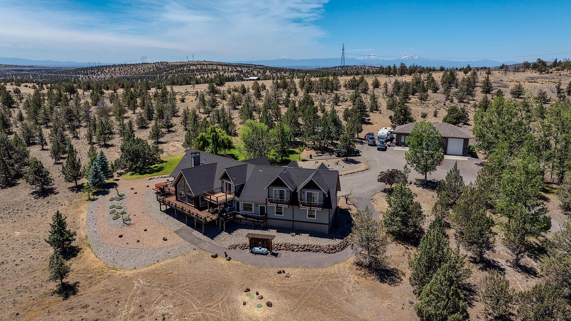 A two-story house with a dark roof in a wooded, arid landscape; curved gravel driveway and parked vehicles nearby among pines and open land in the distance.] ,