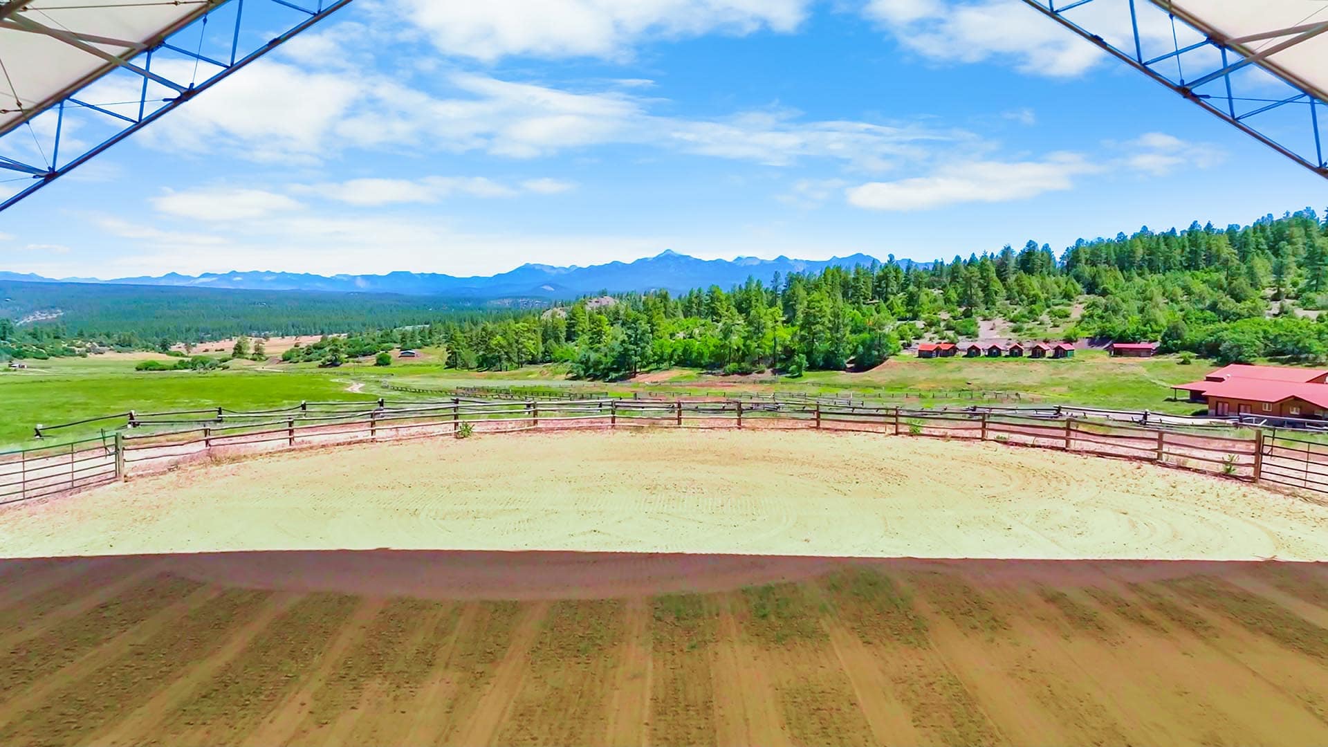 Outdoor horse arena with a wooden fence, sandy footing, green pastures, pine trees, red-roofed buildings, and distant mountains under a blue sky.”,