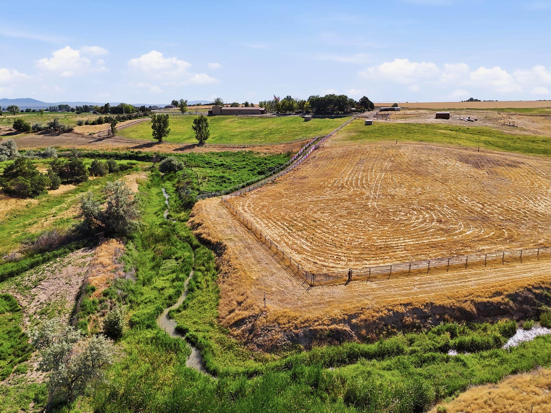 A rural farmland landscape with green fields, a winding ditch, and a long wooden fence curving toward distant buildings under a blue sky.