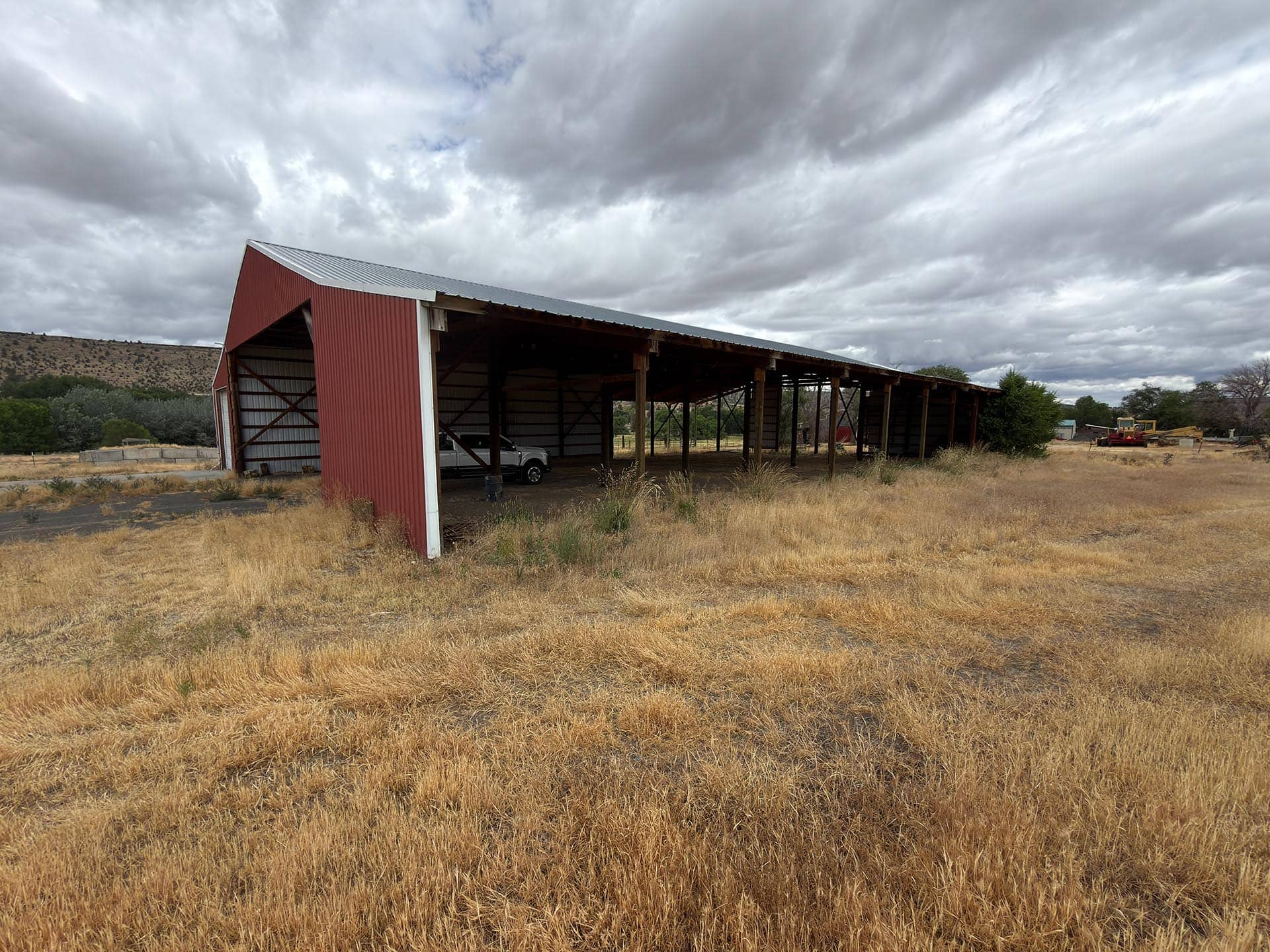 Long red metal barn with open sides in a dry, grassy field under a cloudy sky