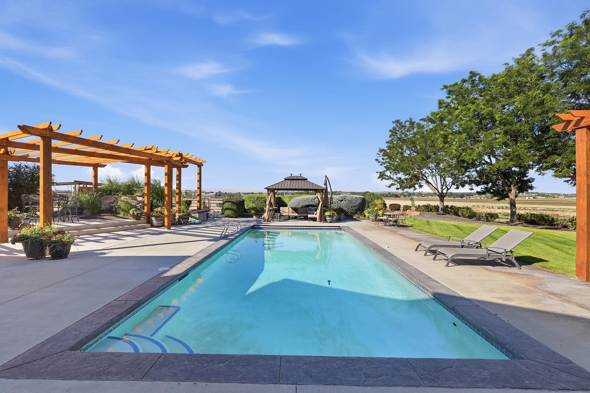 Poolside backyard with a rectangular pool, wooden pergola on the left, lounge chairs, and a seating gazebo against a open landscape with trees.