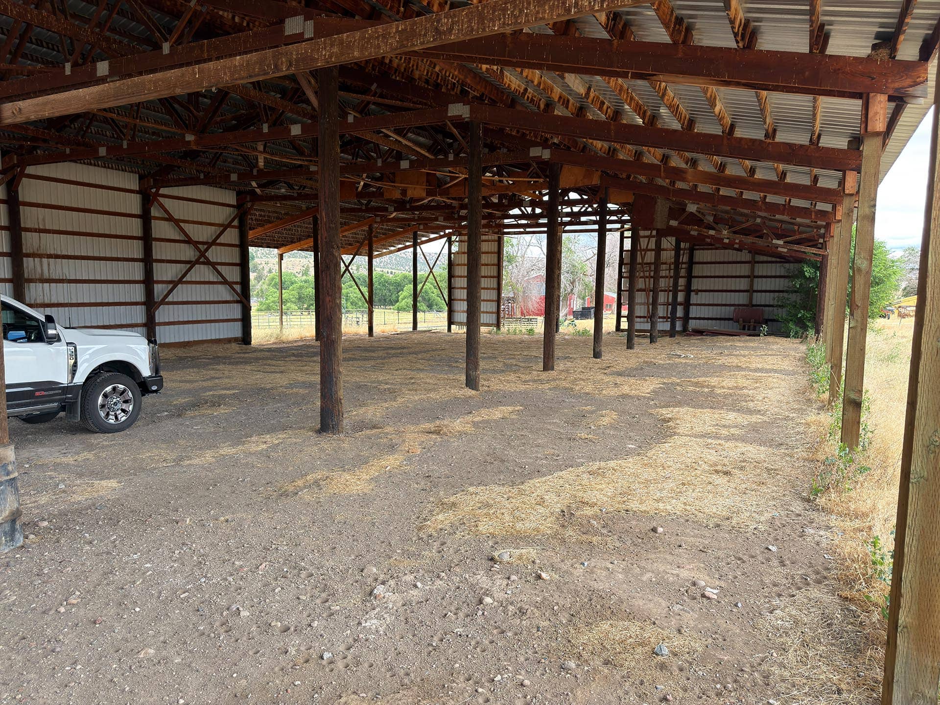 Open-sided wooden barn with exposed beams and dirt ground; a white pickup parked on the left.