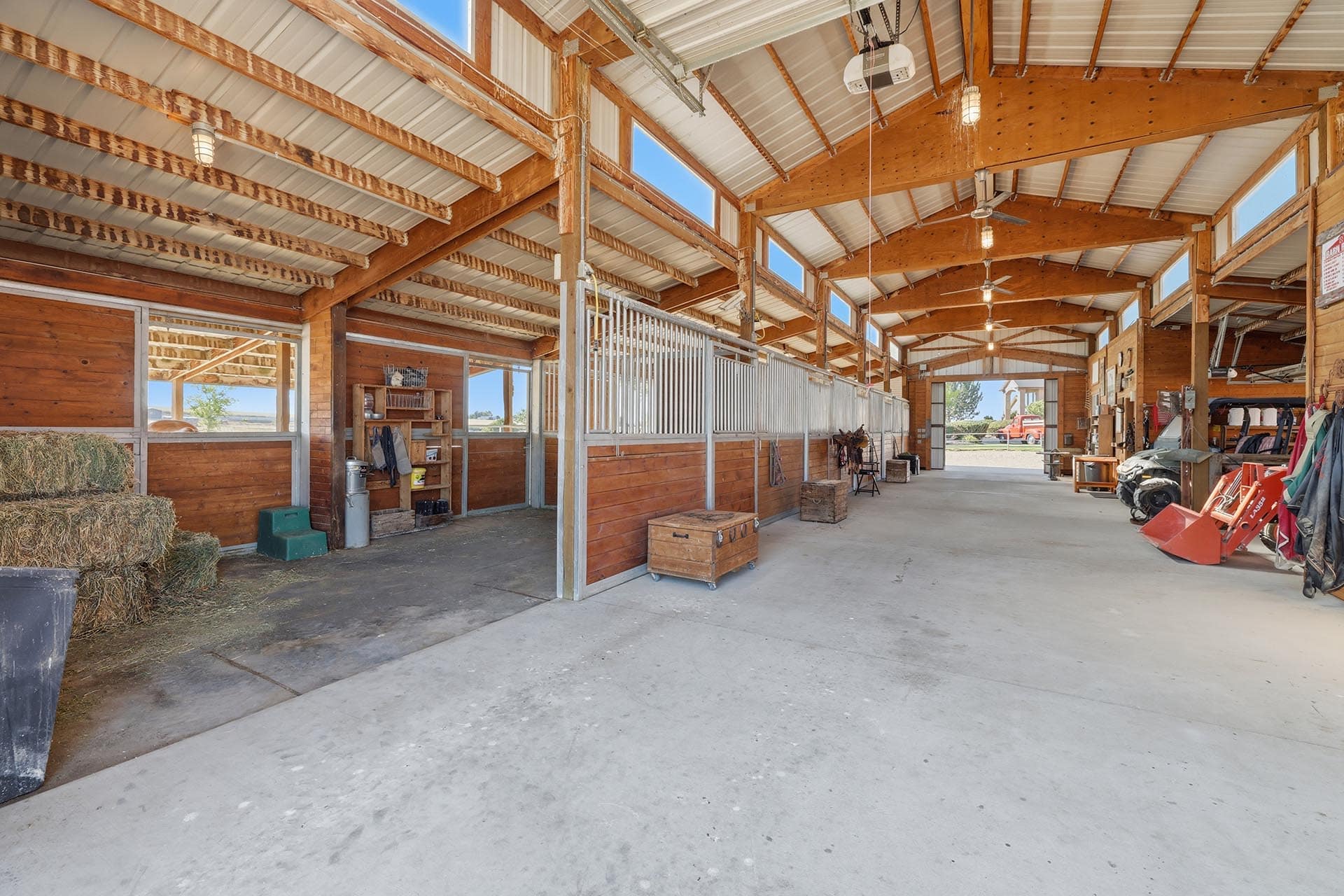 Interior of a neat horse barn with wooden stalls, hay bales, and farm gear along the walls; wide concrete aisle and open doorway to outside.