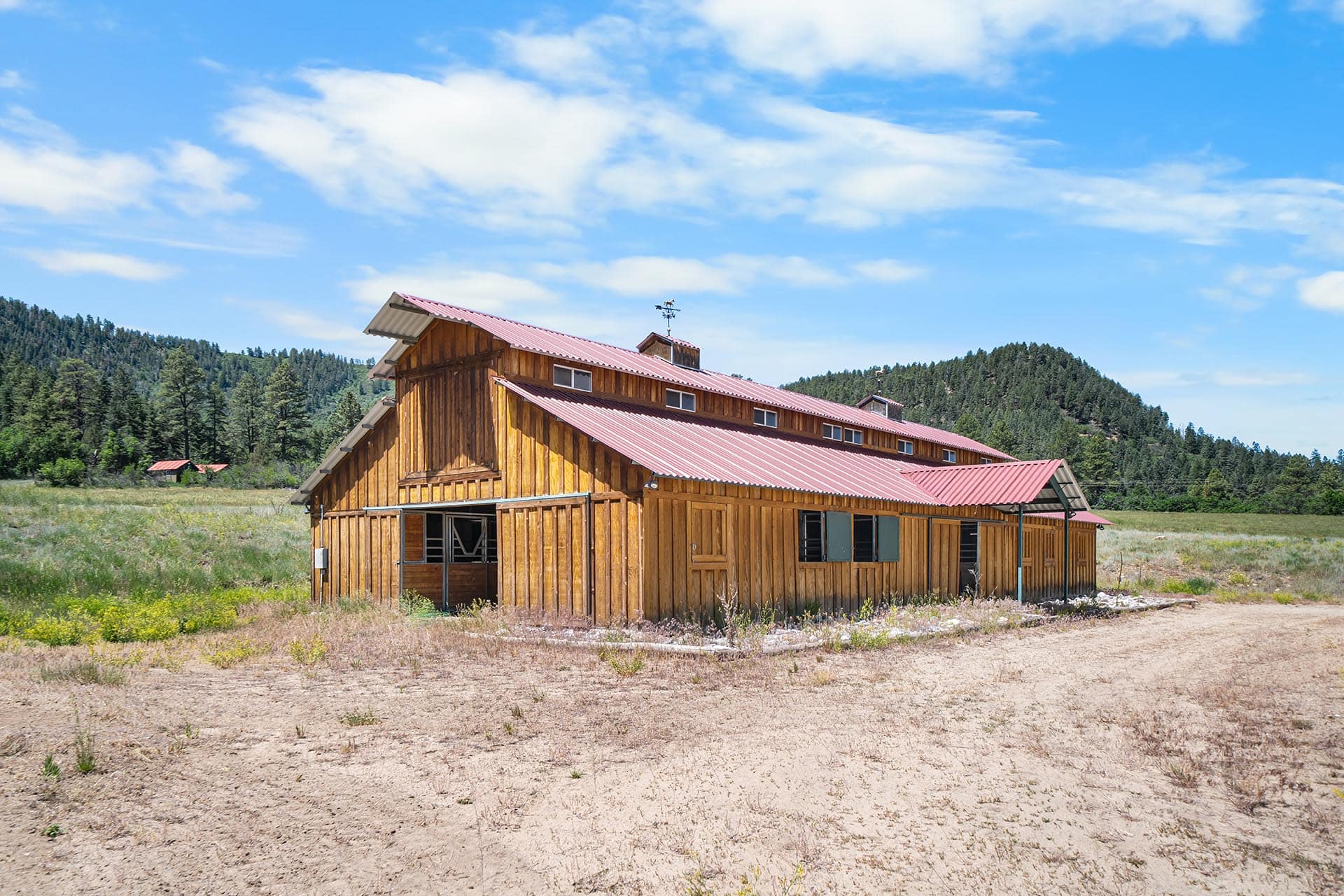 Wooden barn with a red metal roof in a grassy field, with pine trees and hills under a blue sky.