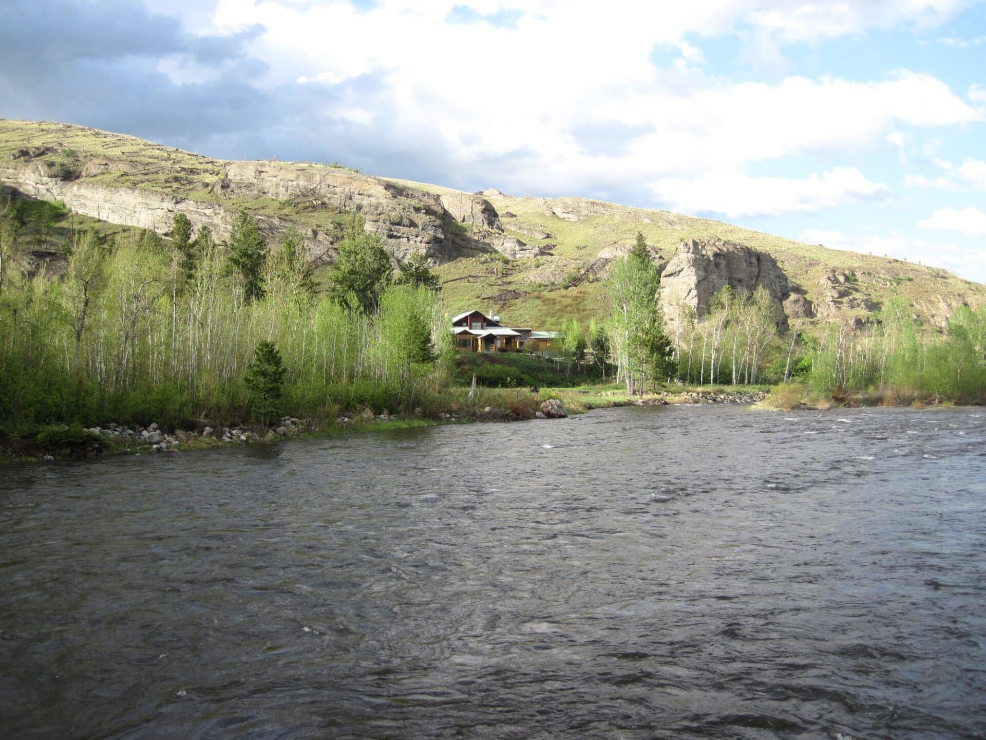 A wooden cabin sits by a river at the base of a rocky hillside, surrounded by green trees and a blue sky with clouds.