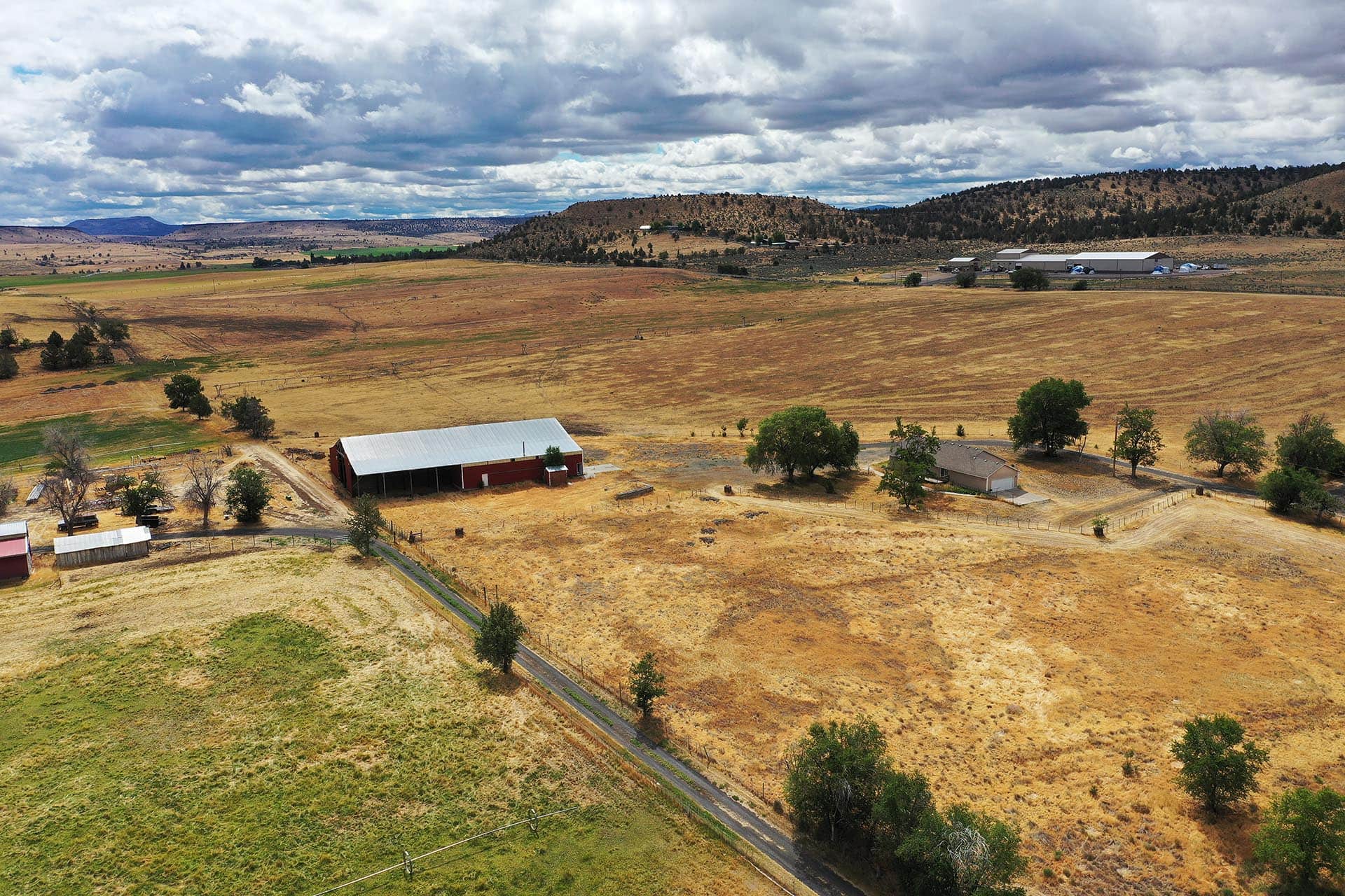 Rural farm landscape with a red barn, long road, and dry golden fields under a cloudy sky.