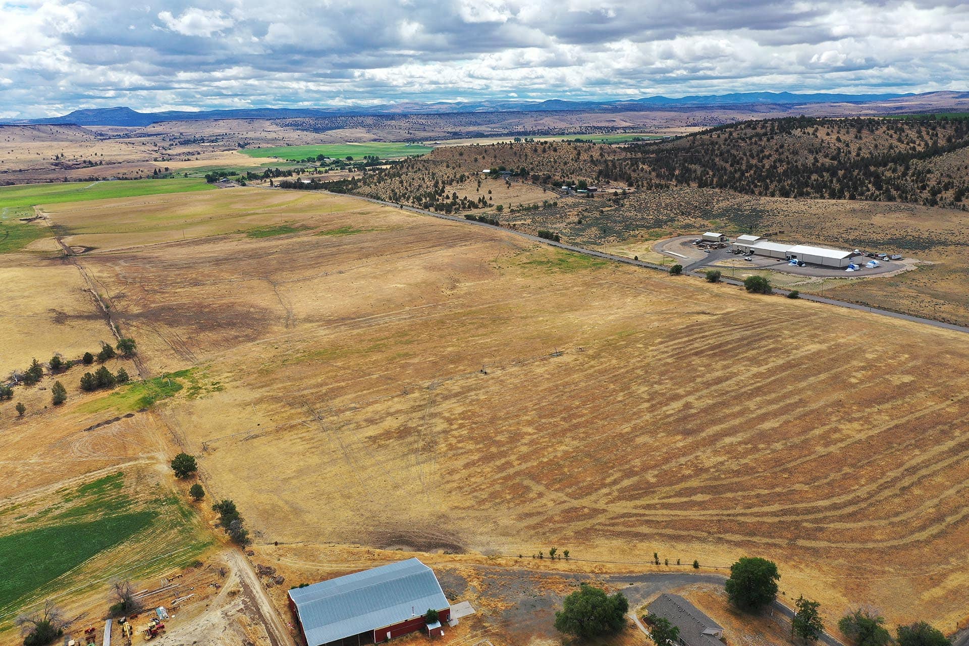 Aerial view of rural farmland with brown fields, green patches, and a few farm buildings along a curved road.